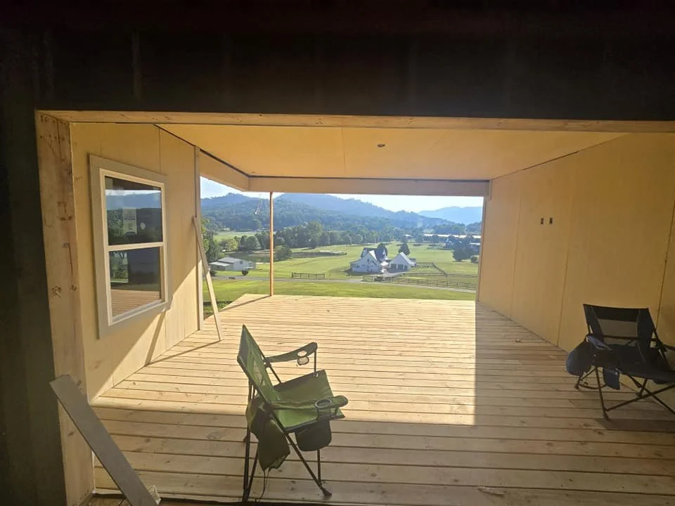 View from inside a house under construction, looking out onto a porch with chairs and a rural landscape with houses, trees, fences, and mountains in the distance.