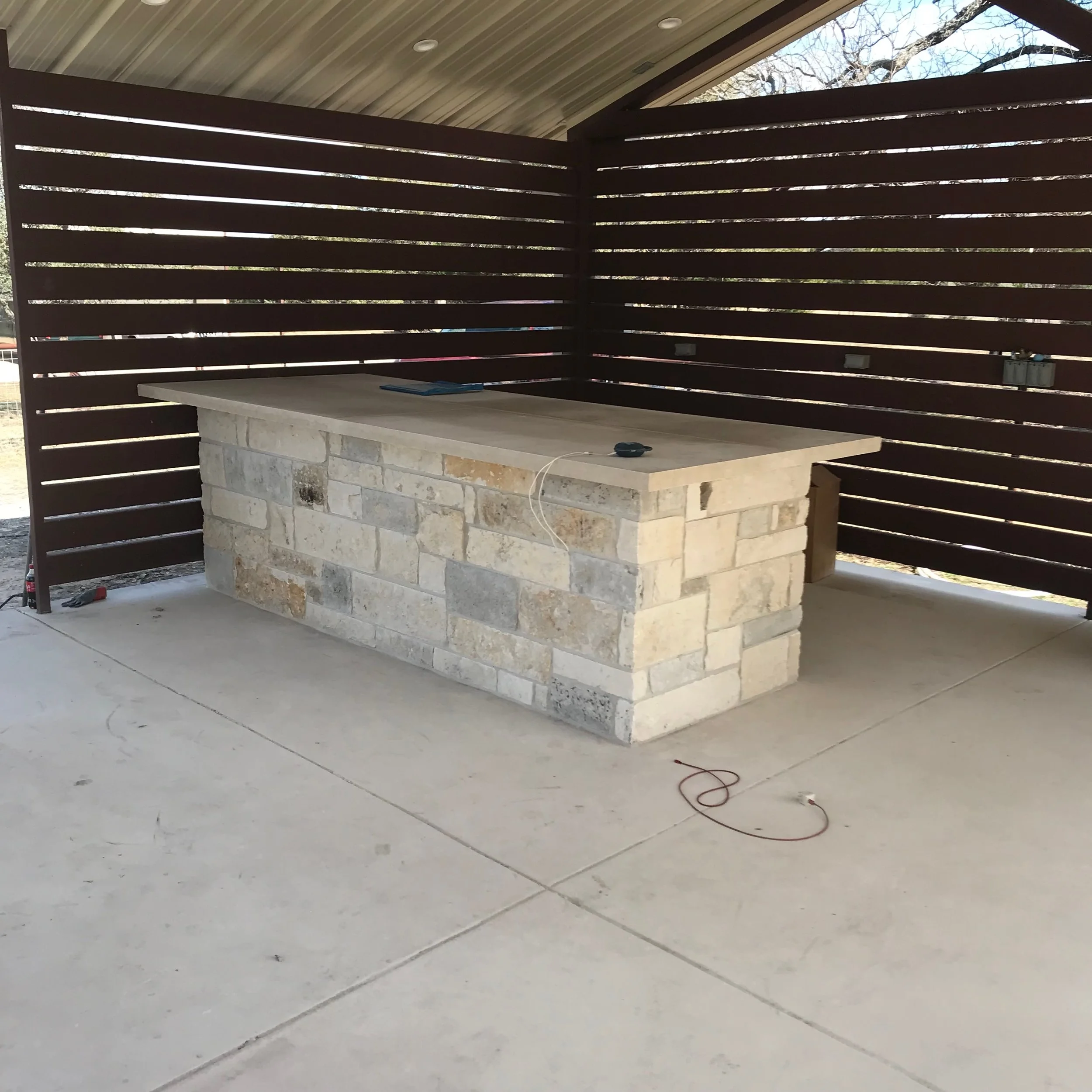 Outdoor stone bar with a concrete countertop, surrounded by a wooden slatted privacy fence, and a concrete floor.
