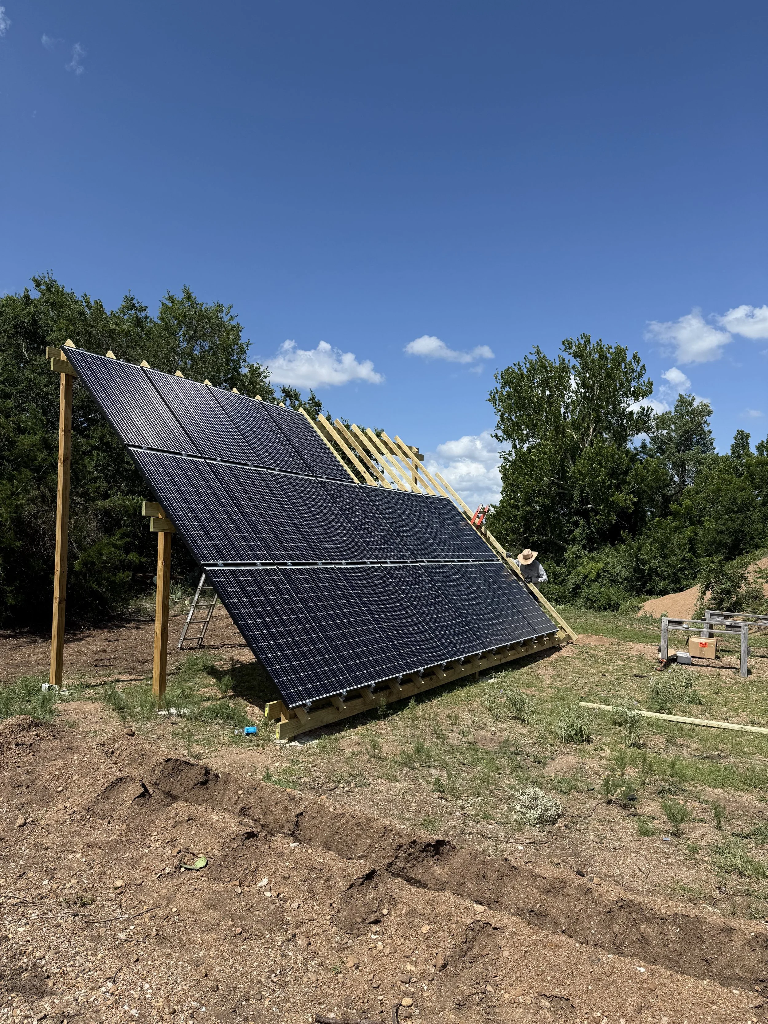 Solar panels under construction on a rooftop with ladders, wood framing, and a worker installing, surrounded by trees on a clear, sunny day