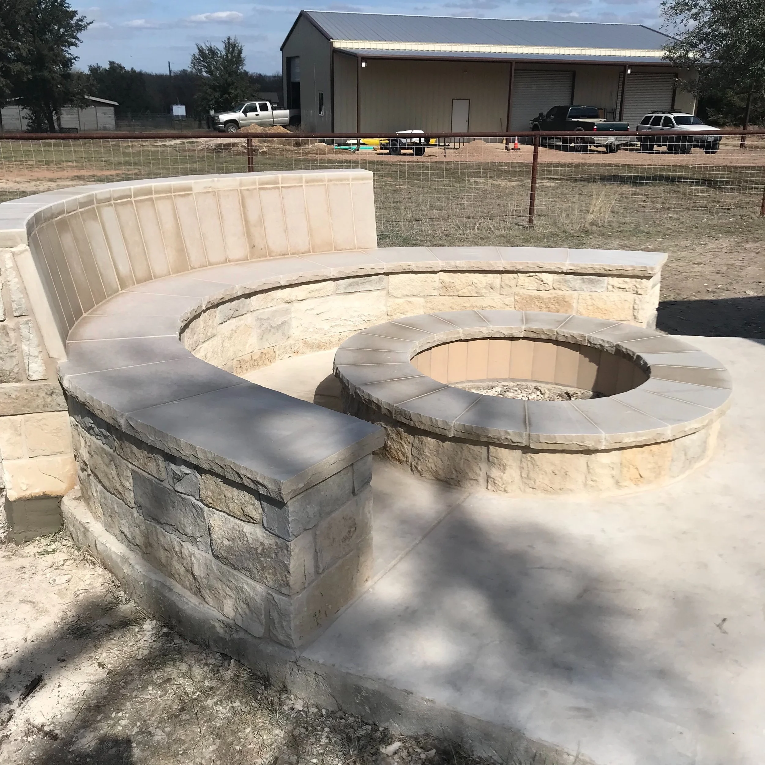 Concrete fire pit with surrounding stone and tiled bench, located outdoors in a yard under a partly cloudy sky.