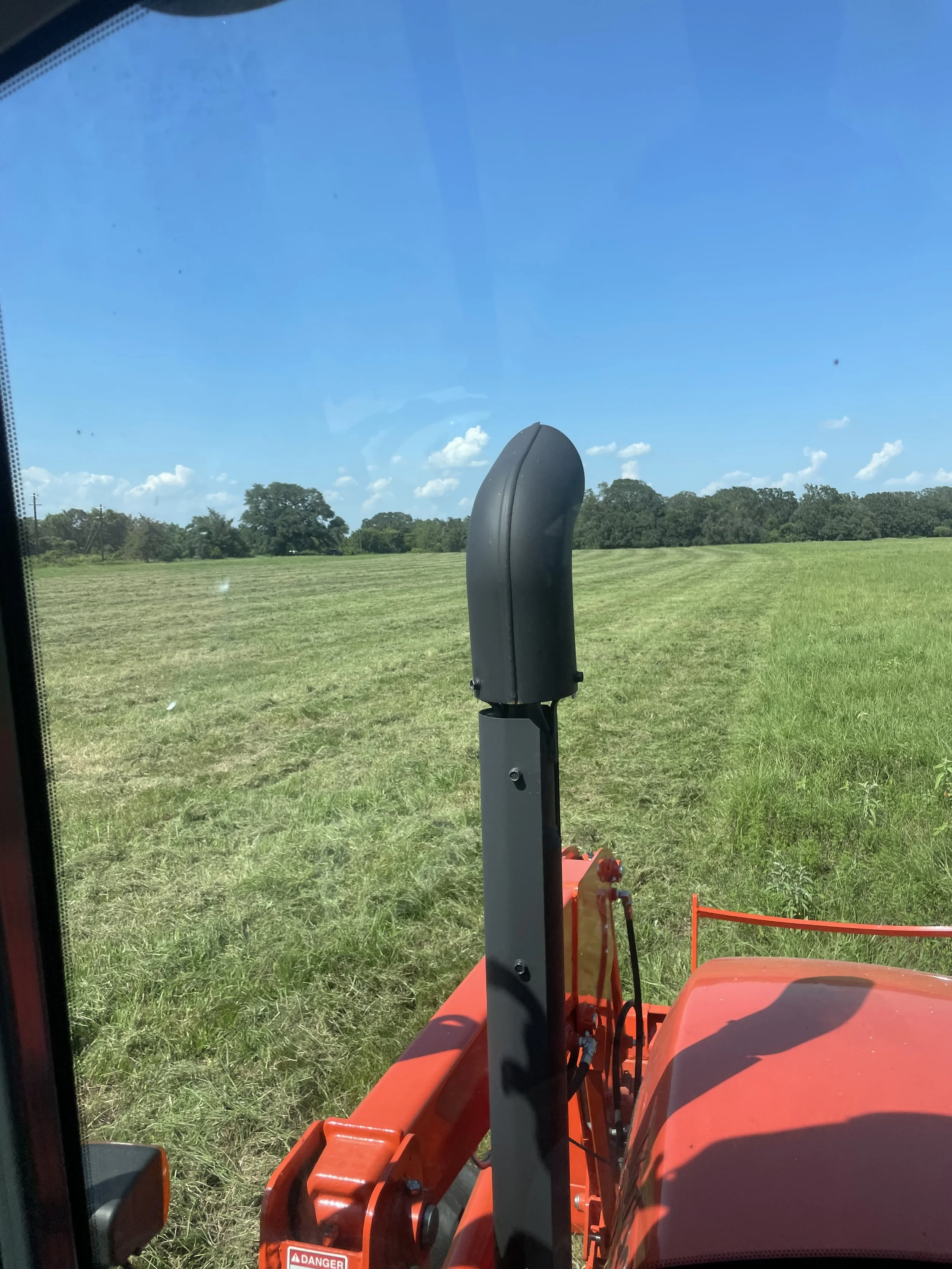 View from inside a tractor cabin looking out at a green field with blue sky and some clouds, showing part of the tractor's black exhaust pipe and orange hood.