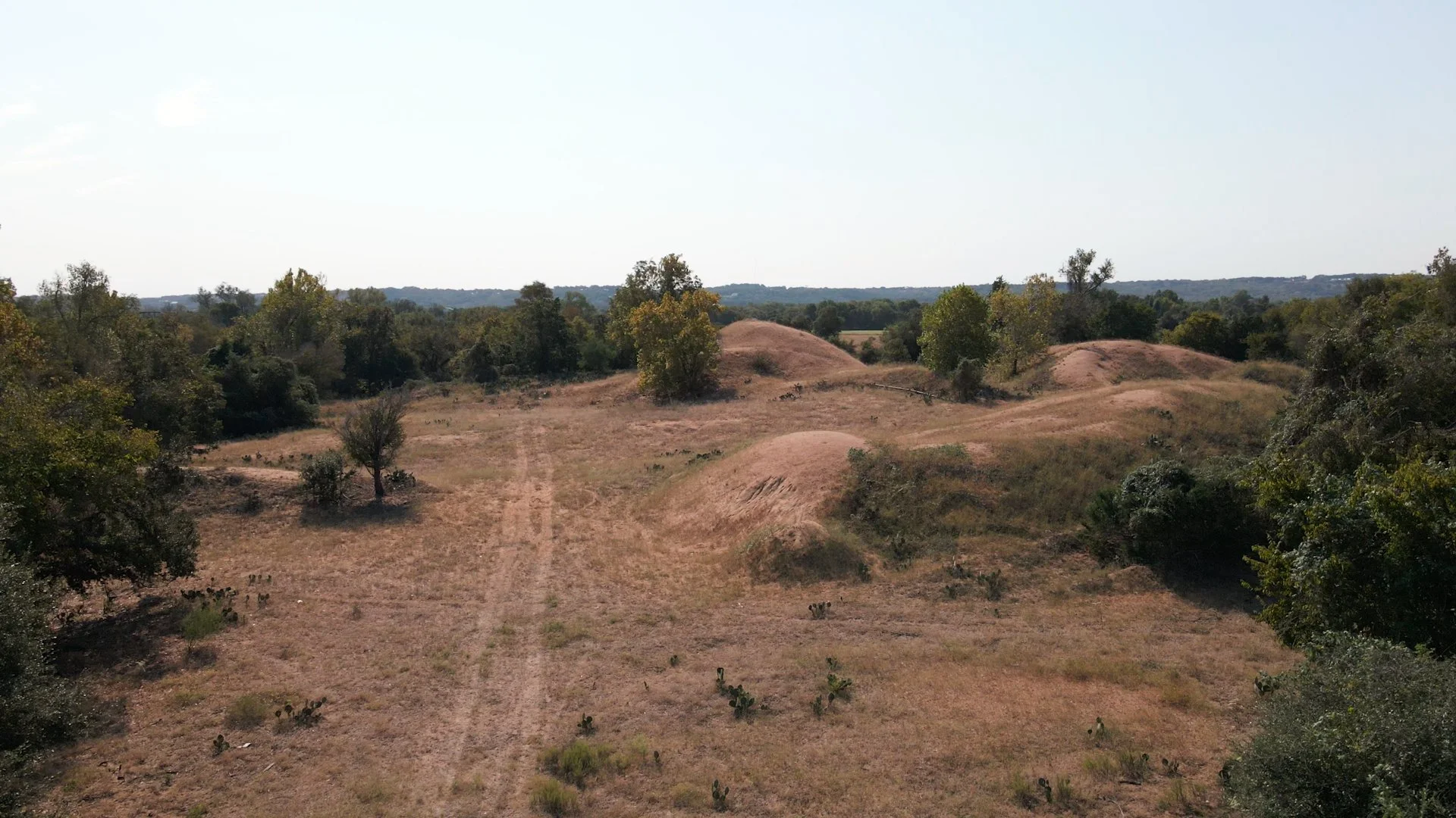 A dry landscape with rolling hills, sparse trees, and a dirt path, under a clear sky.