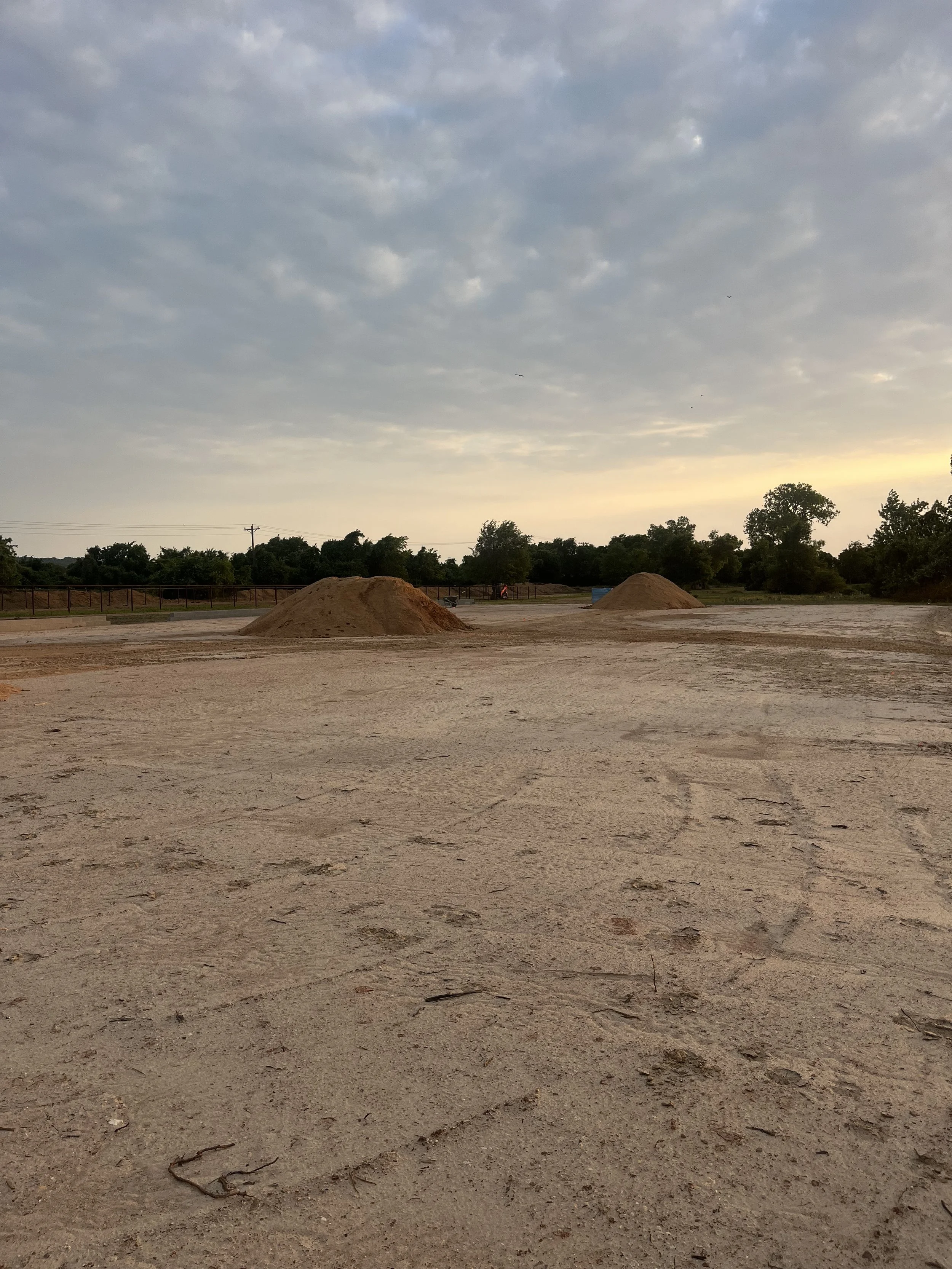 A dirt lot with two small piles of sand or dirt, trees and a cloudy sky in the background during sunset.