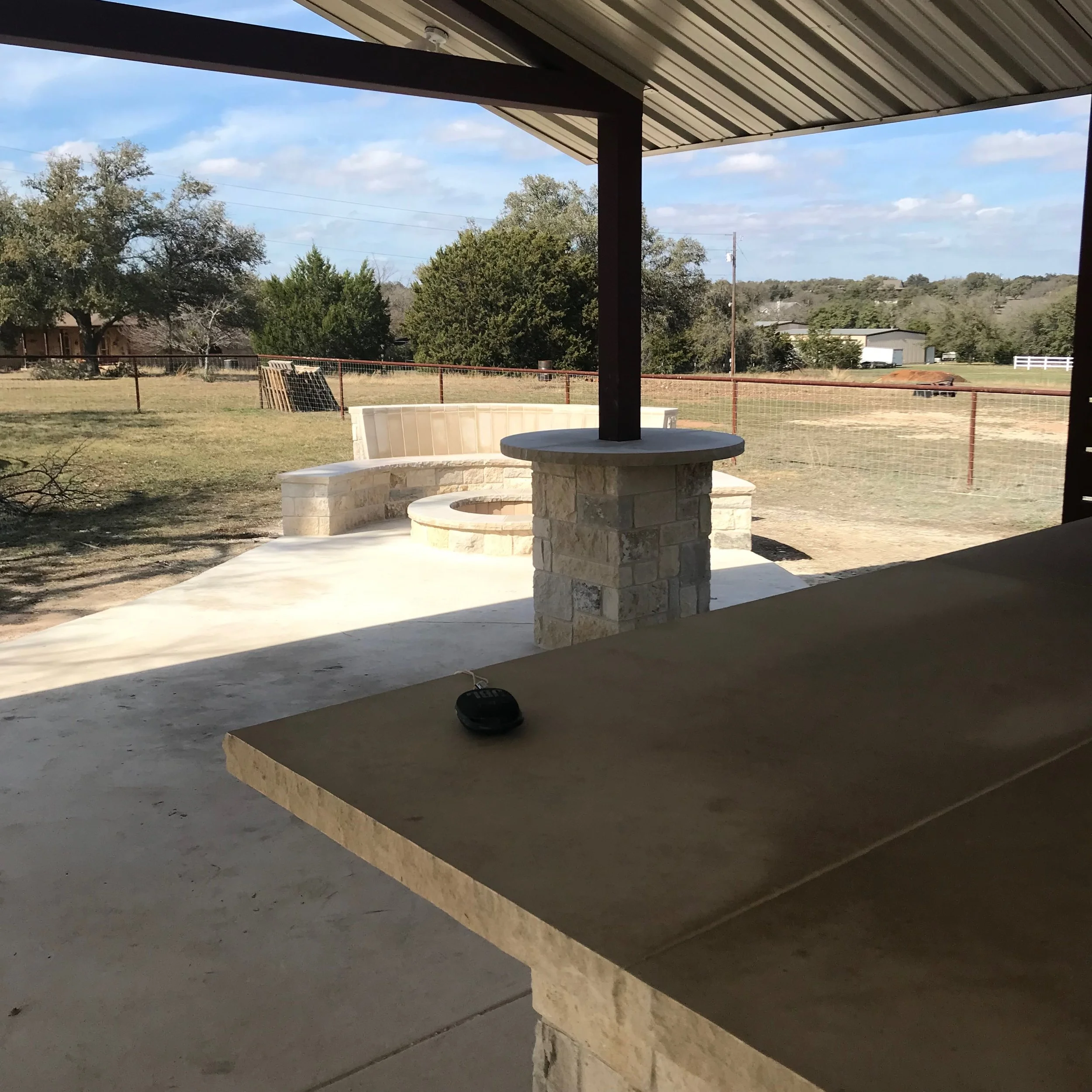 View from a covered patio showing a stone fire pit with a circular seating area in a backyard, enclosed by a brown metal fence, with trees and buildings in the distance under a partly cloudy sky.