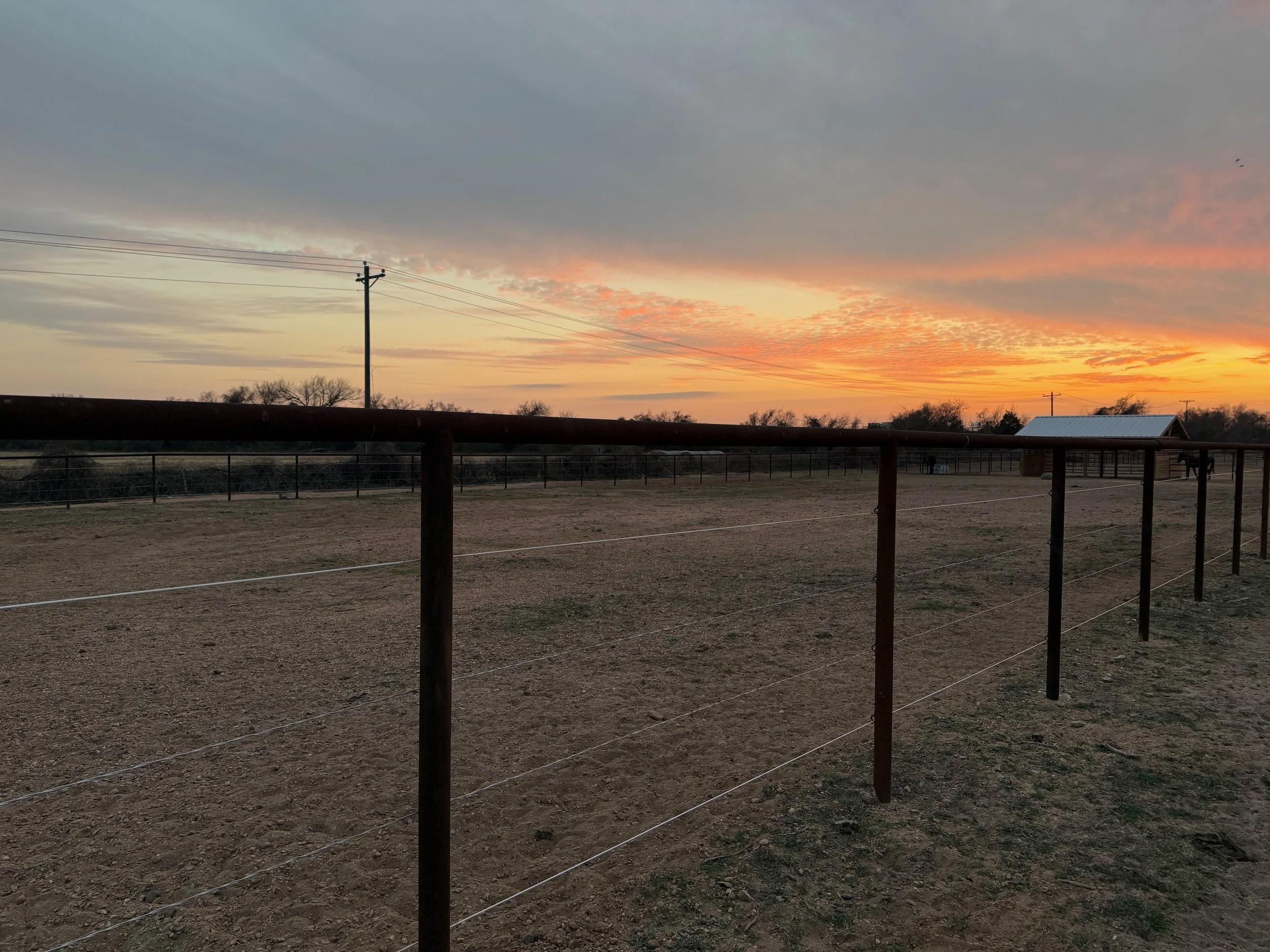 Fenced dirt field at sunset with a small building and power lines in the distance.