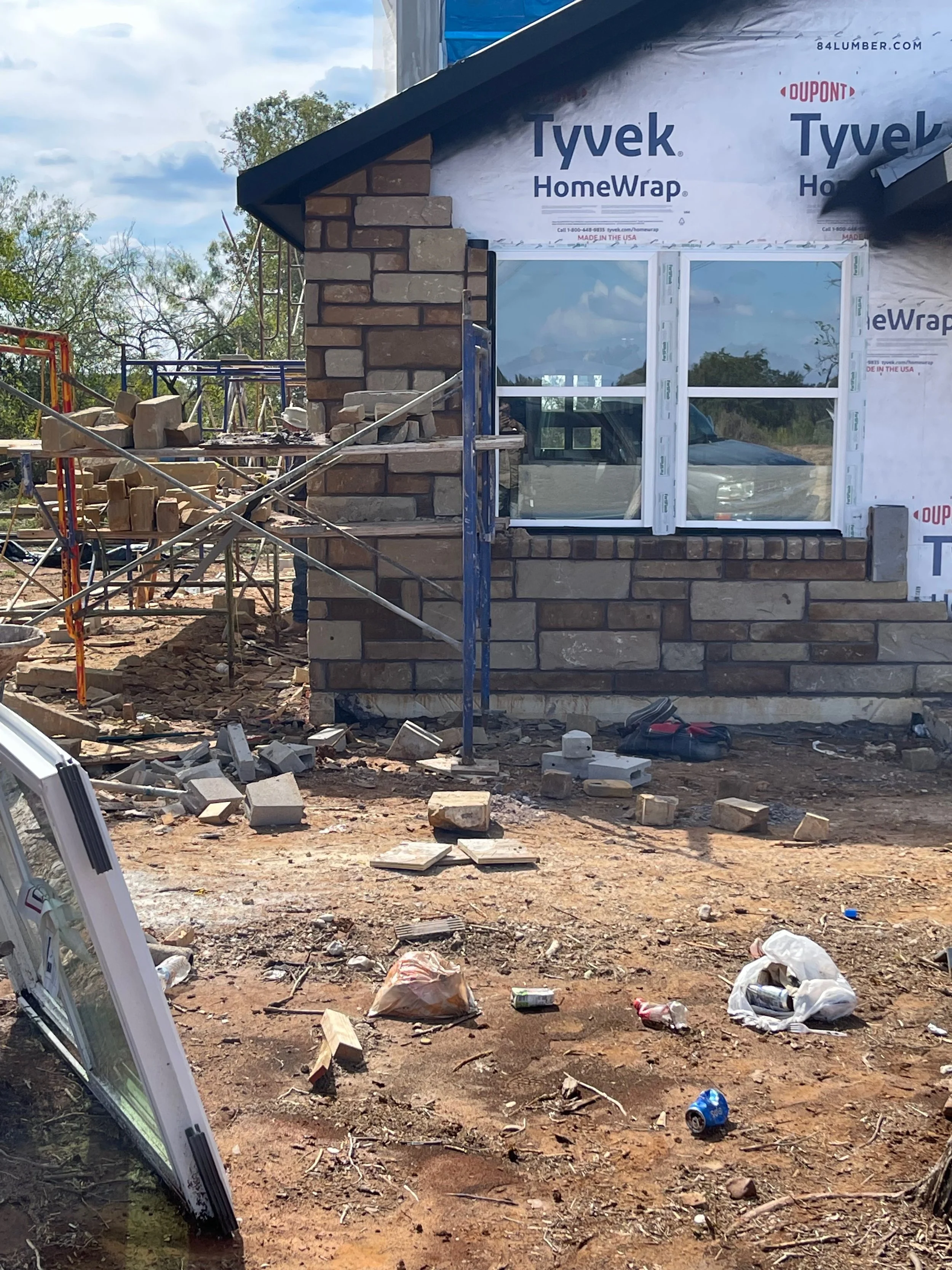 Construction site with brick wall and window, construction debris and tools scattered on dirt ground.