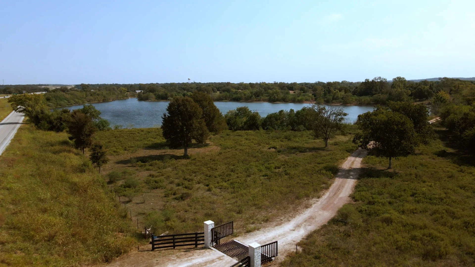 Aerial view of a landscape with a dirt road leading to a lake, surrounded by trees and open fields under a blue sky.