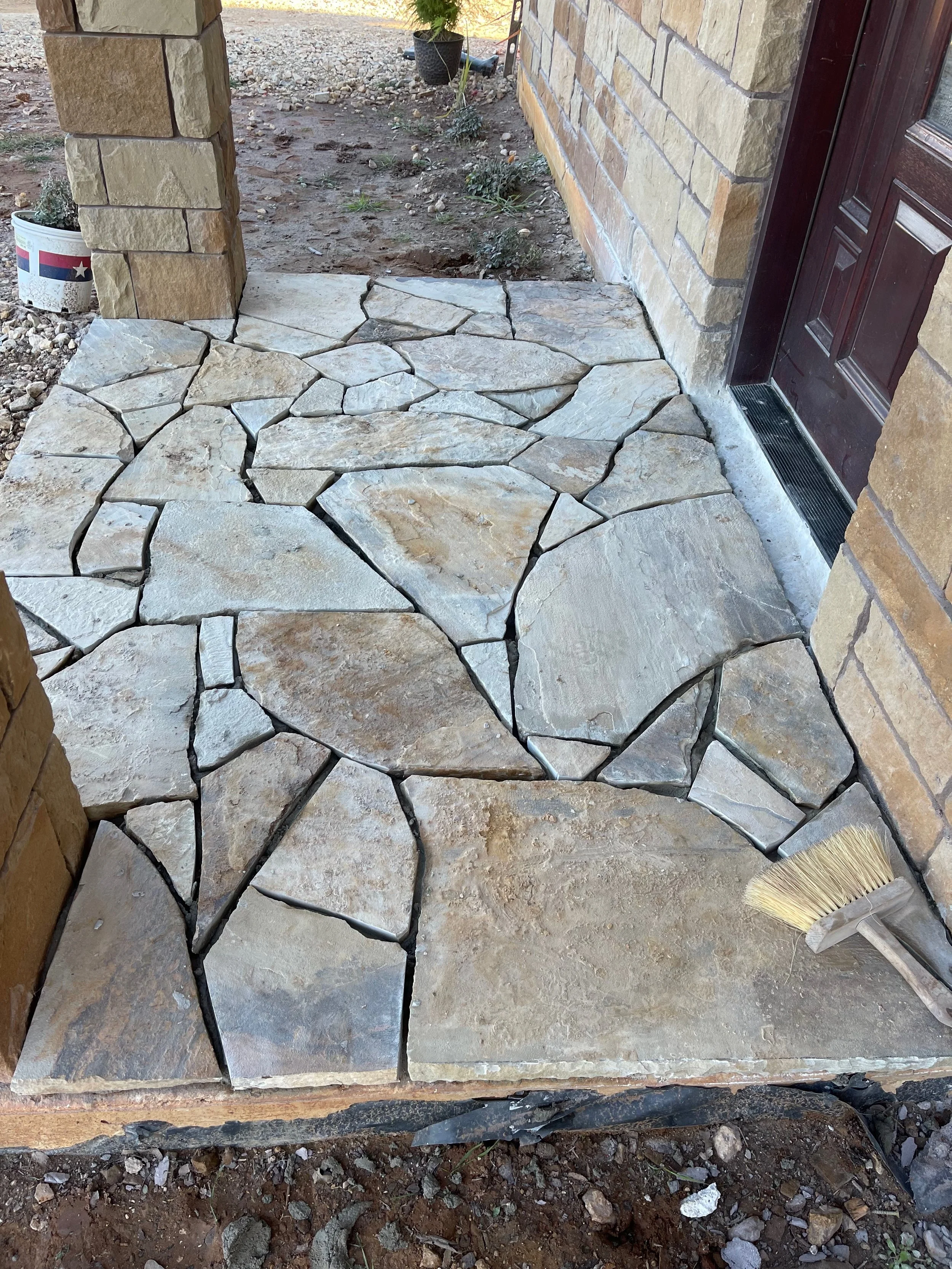 Stone porch with unevenly placed flagstone slabs, a broom resting on the edge, and a brick wall and door nearby.