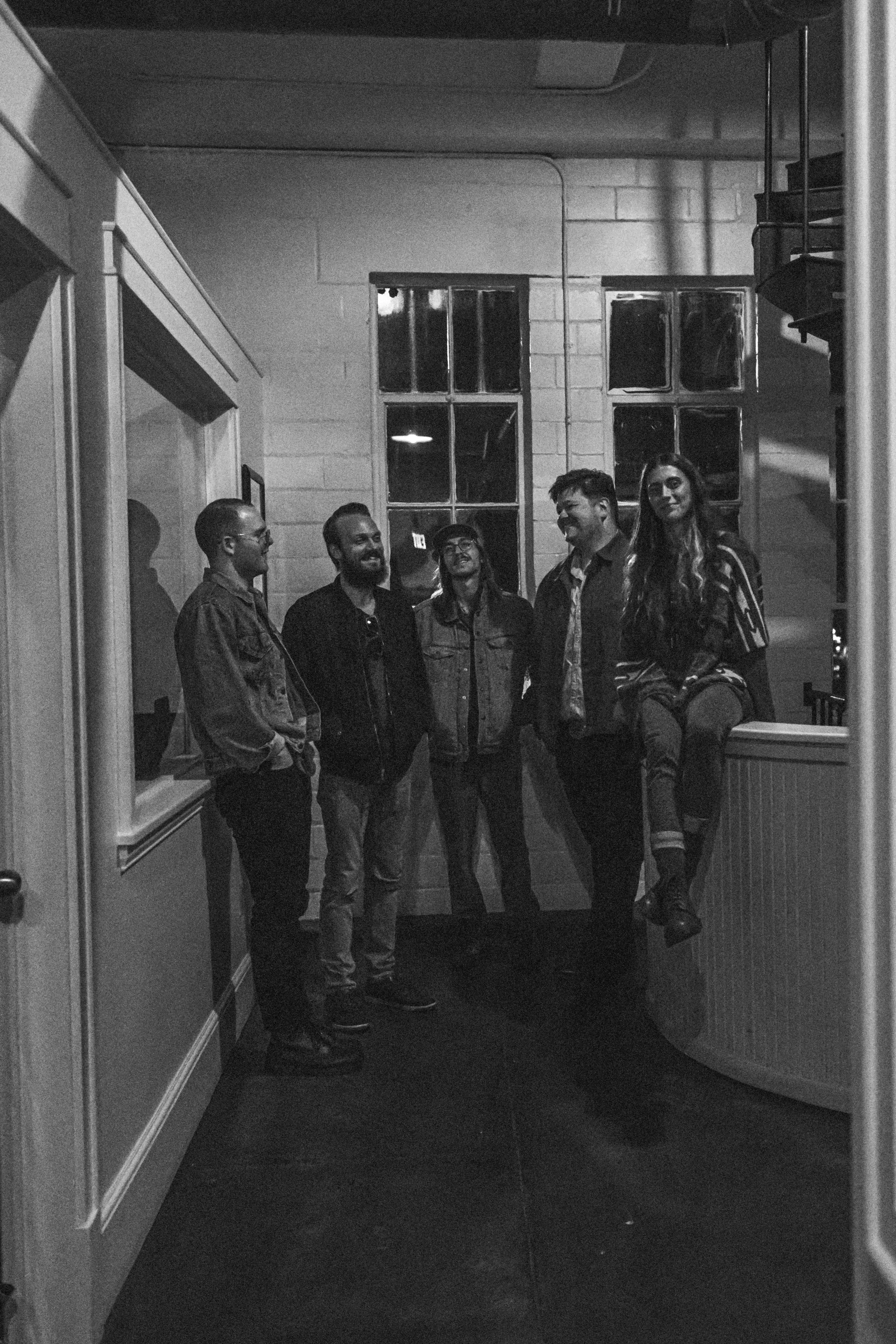 Group of five people standing and sitting in a dimly lit room with large windows, high ceilings, and a spiral staircase. All are smiling or laughing.