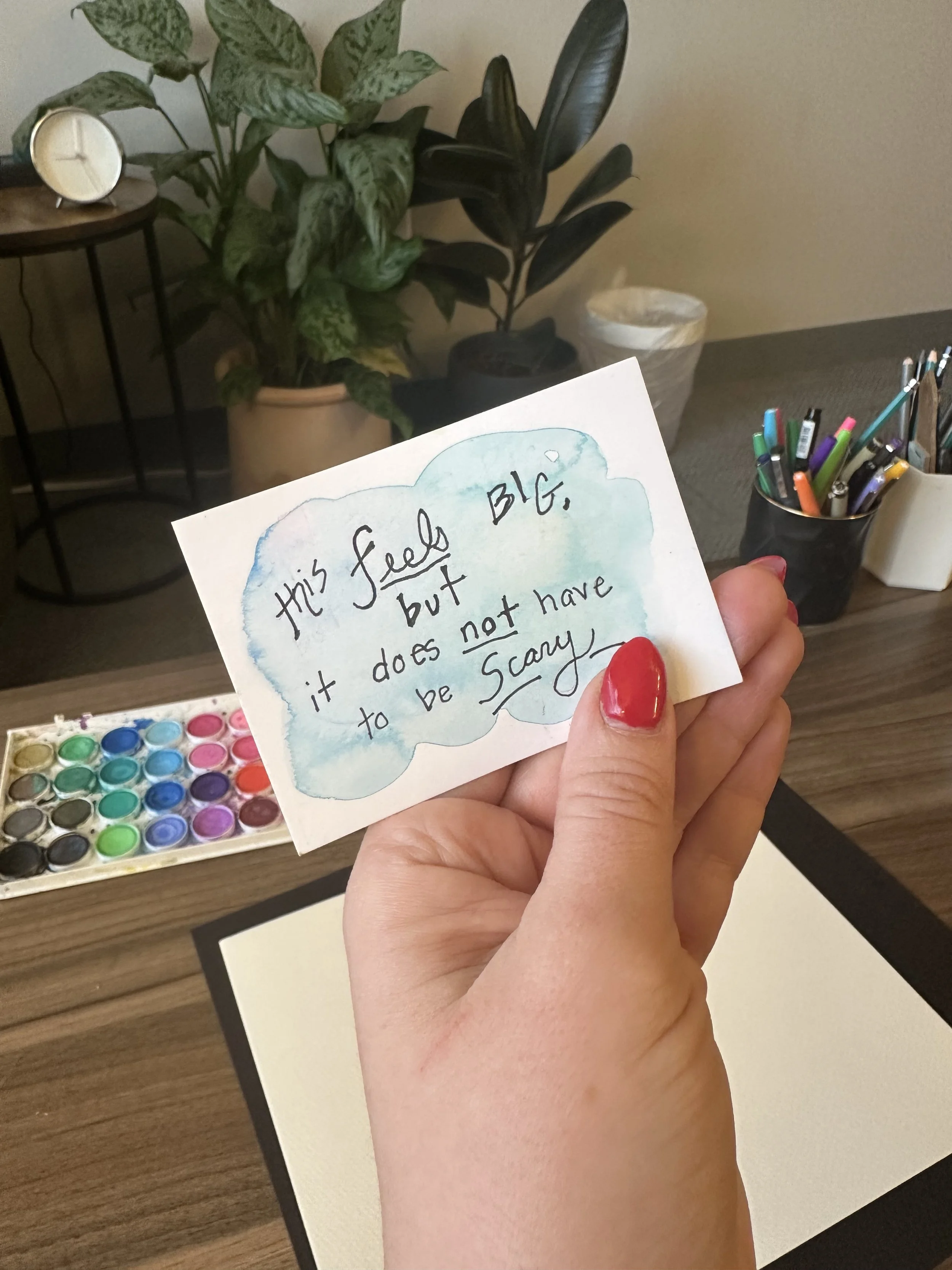 A hand holds a note saying, "this feels big but it does not have to be scary" over a desk with art supplies.