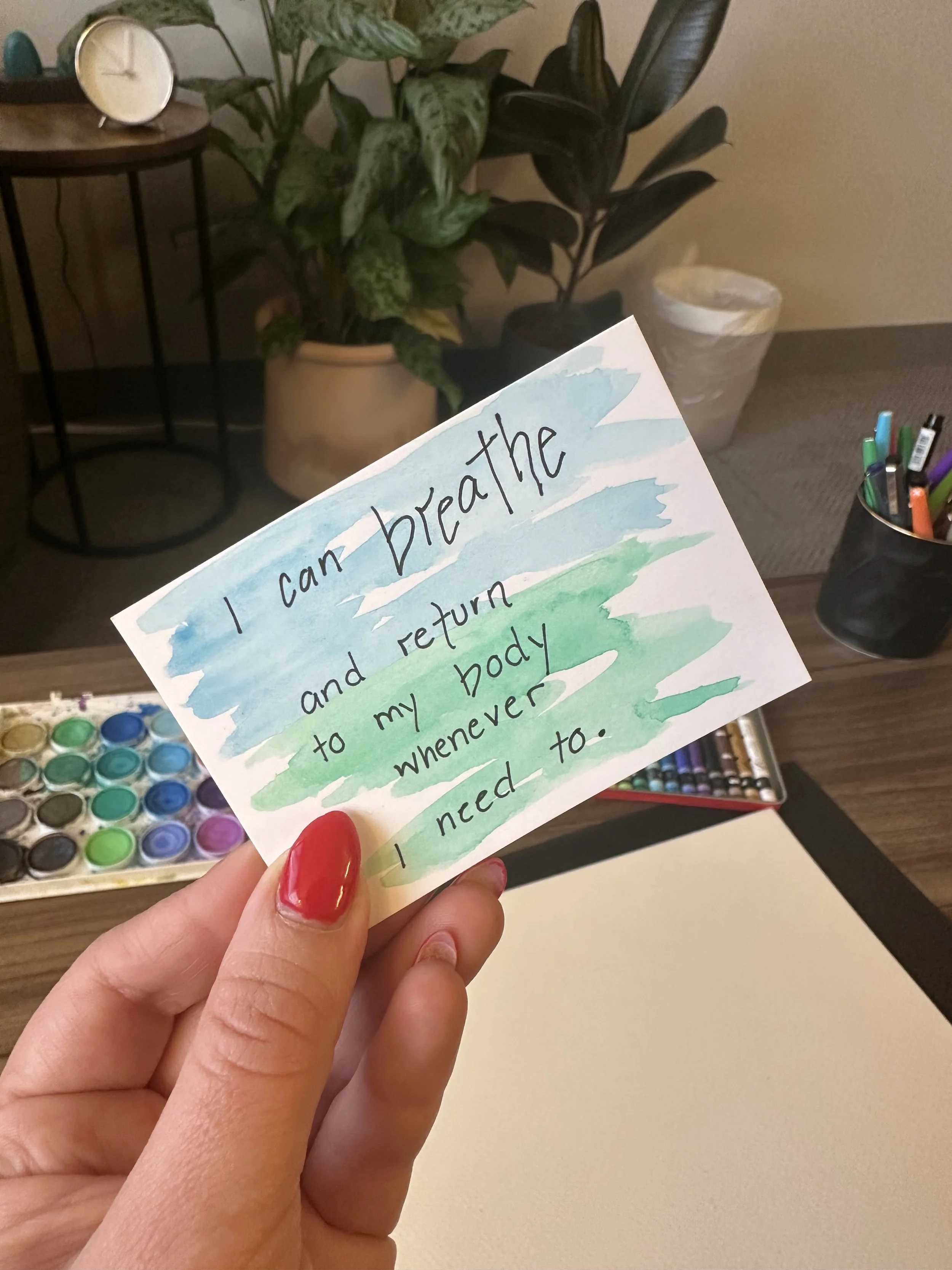 A hand holds a note saying, I can breathe and return to my body whenever I need to, over a desk with art supplies.