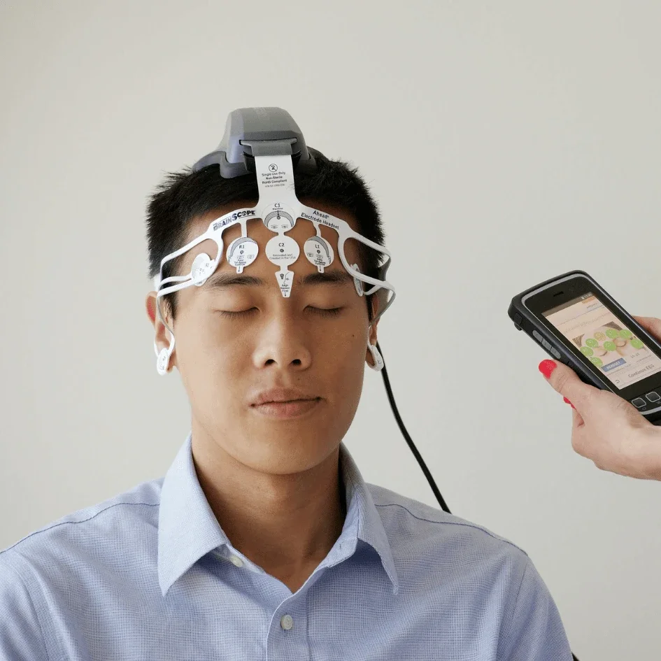 Man participating in an EEG test, wearing a cap with electrodes on his head while a person operates a mobile device nearby.