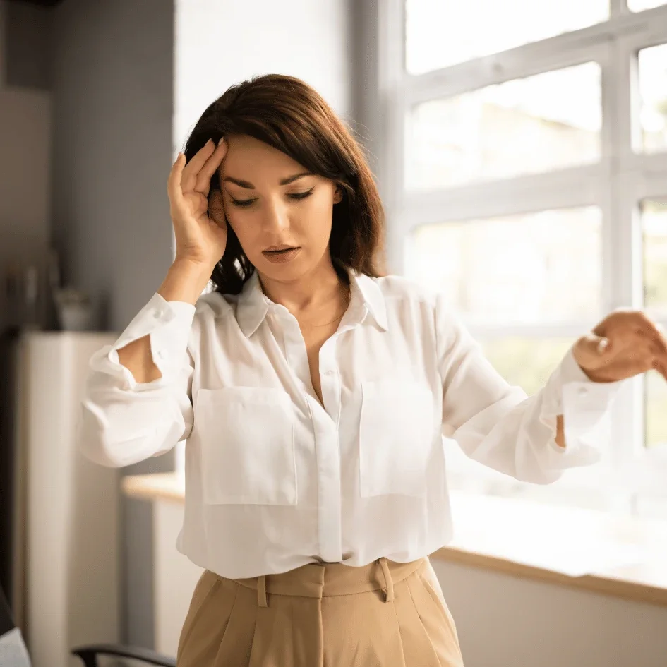 A young woman in a white blouse and beige trousers is holding her forehead with one hand, appearing worried or stressed, in a bright room with large windows.