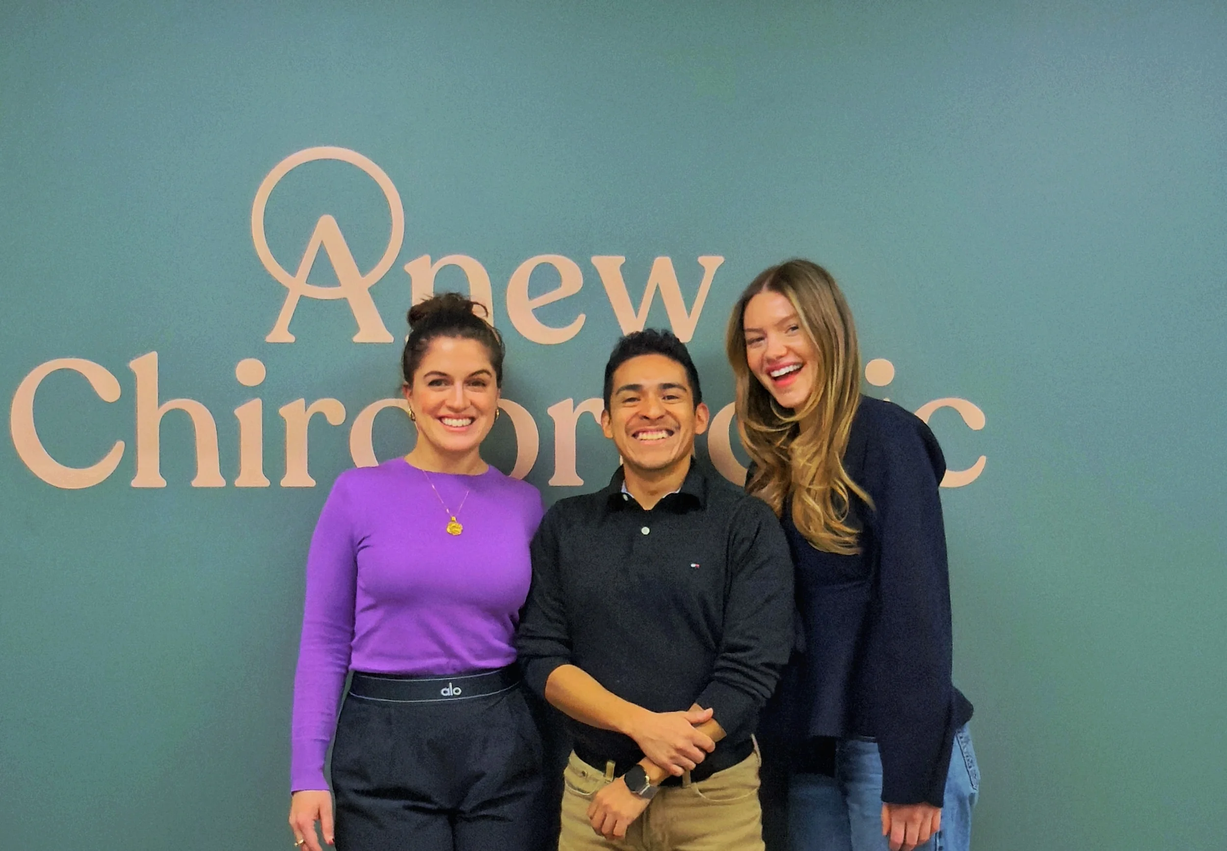 Three people standing together in front of a teal wall with the words 'Anew Chiropractic' on it. The woman on the left has dark hair tied up and is wearing a purple top. The man in the middle has dark hair, a big smile, and is wearing a dark shirt. The woman on the right has long blonde hair and is wearing a dark blazer, smiling with her head slightly tilted.