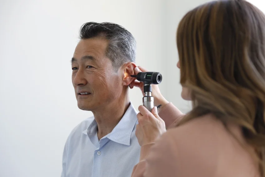 Woman examining middle-aged man's ear with an otoscope in a clinical setting.