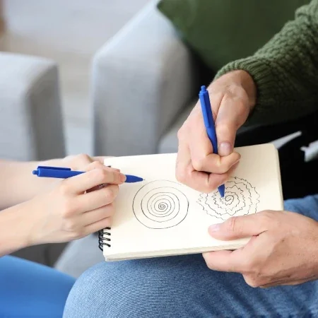 Two people drawing spirals with blue pens on a spiral-bound notebook indoors.