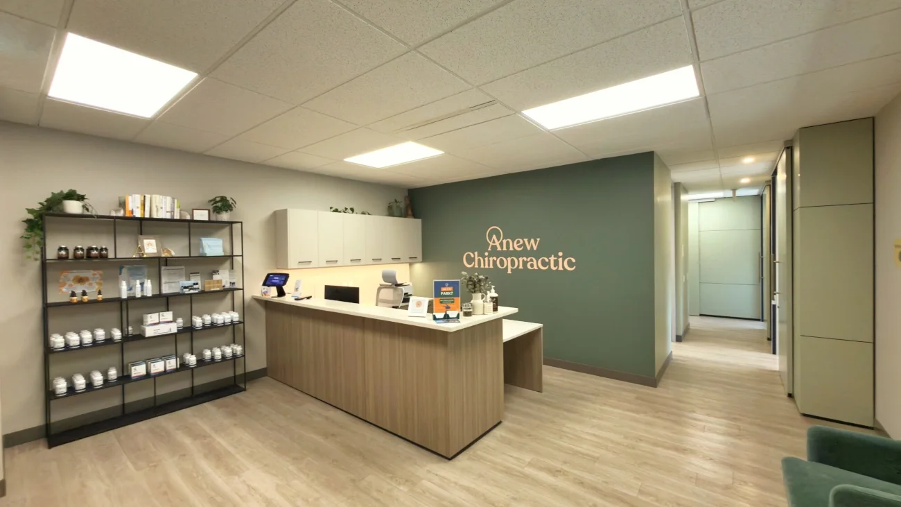Modern chiropractic clinic reception area with a wooden front desk, a shelving unit with supplements, and a green accent wall with the text 'Anew Chiropractic'.