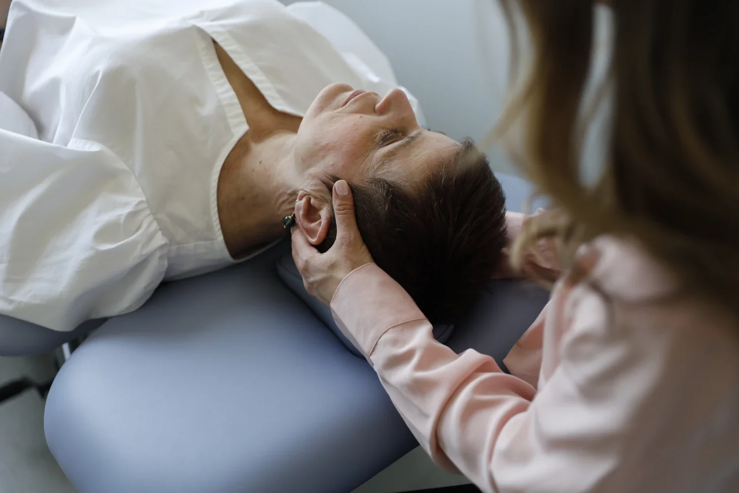 A healthcare professional examines an elderly woman lying on an examination table, touching her head gently.