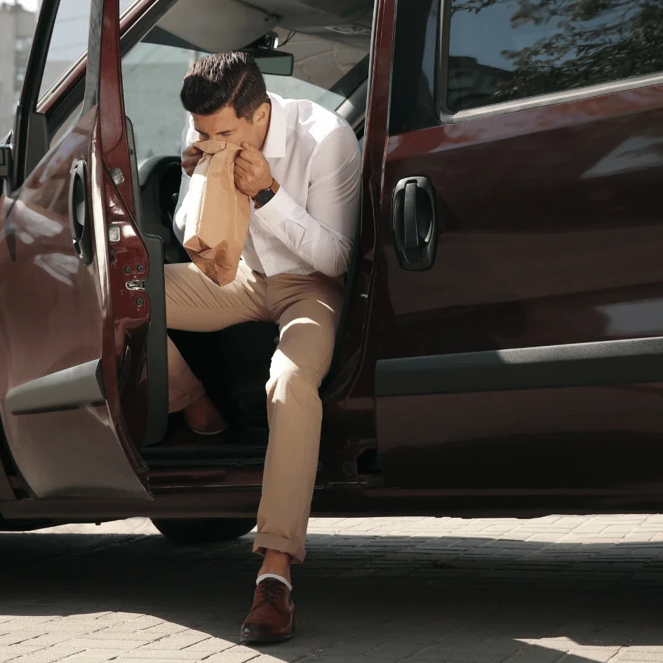 Man sitting in a car door, coughing into a brown paper bag.