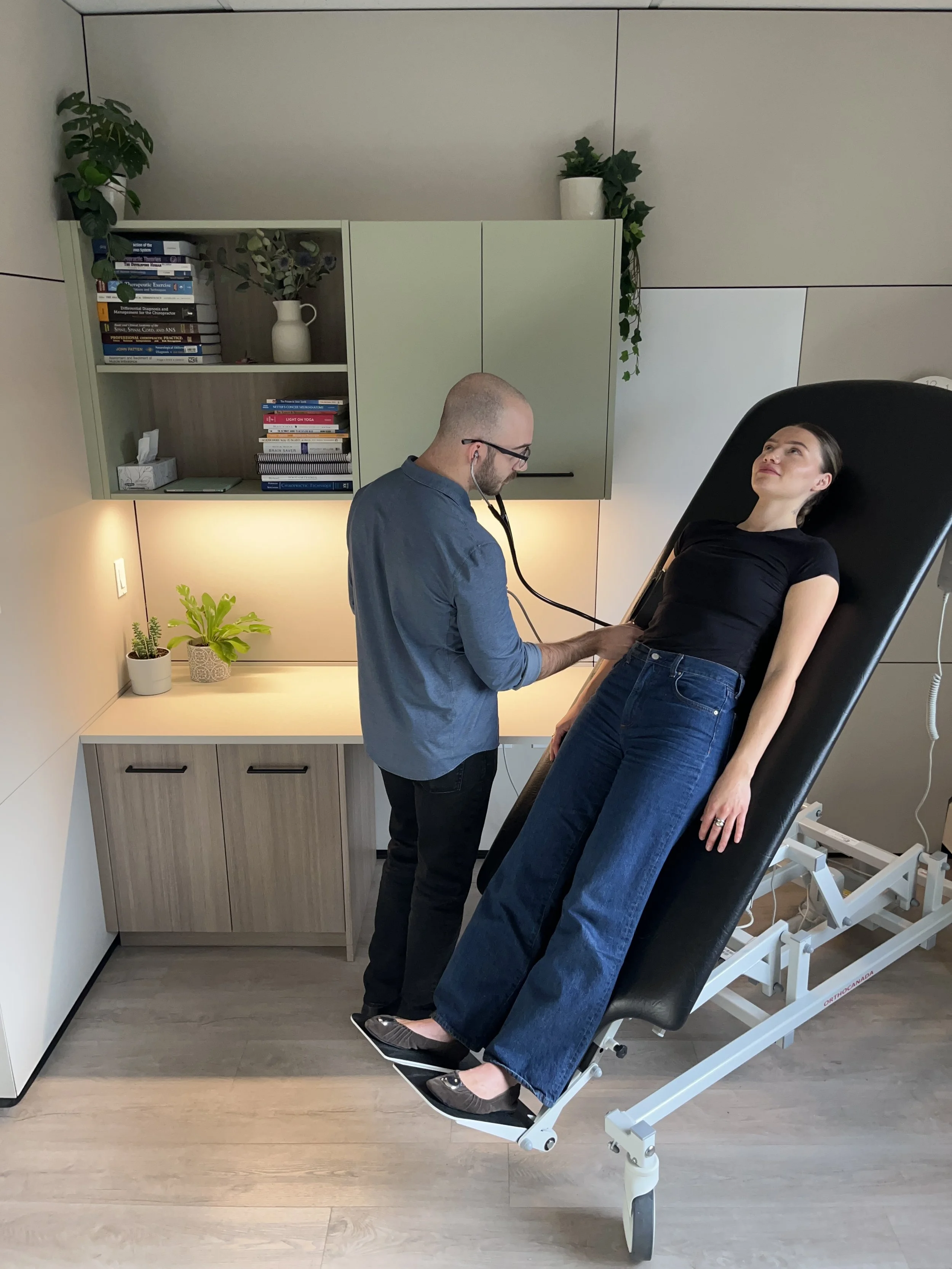 A healthcare professional uses a stethoscope on a woman reclining on a medical examination table in a clinic or medical office. The room has a wooden cabinet with books and plants on top, and a plant on a shelf in the background.