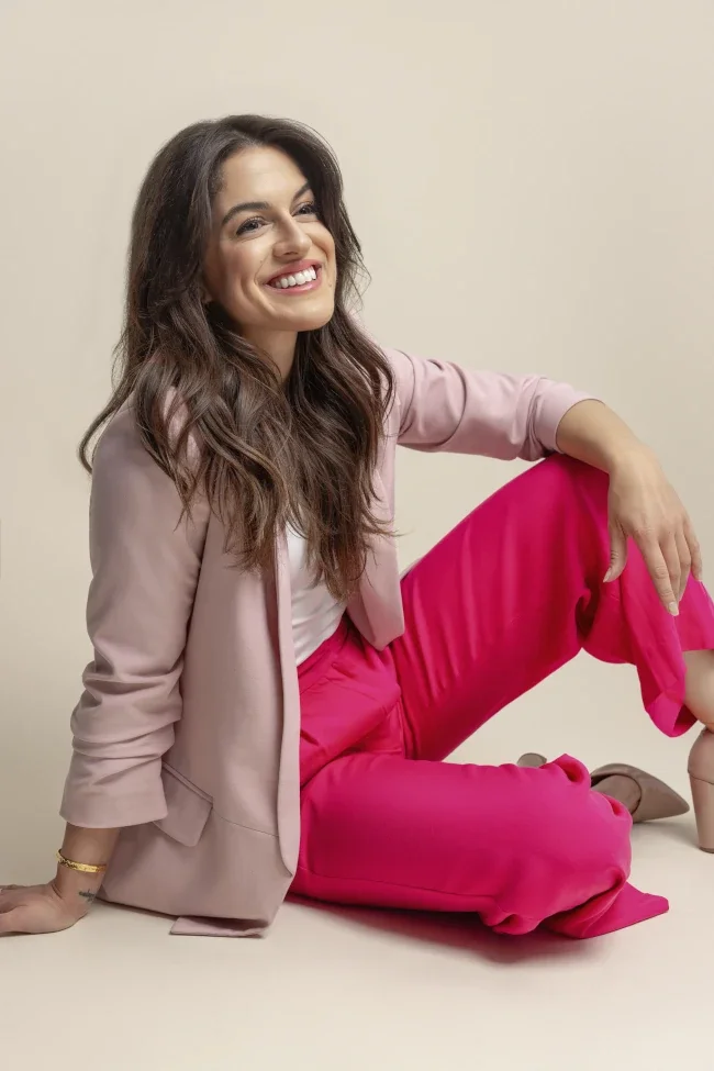 A woman with long brown hair sitting on the floor against a plain light-colored background, smiling, wearing a light pink blazer, white top, and bright pink pants.