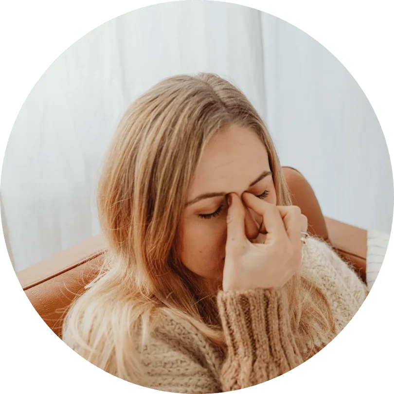 A woman with light brown hair wearing a beige knitted sweater, sitting indoors, with her hand pinching the bridge of her nose and eyes closed, appearing stressed or tired.