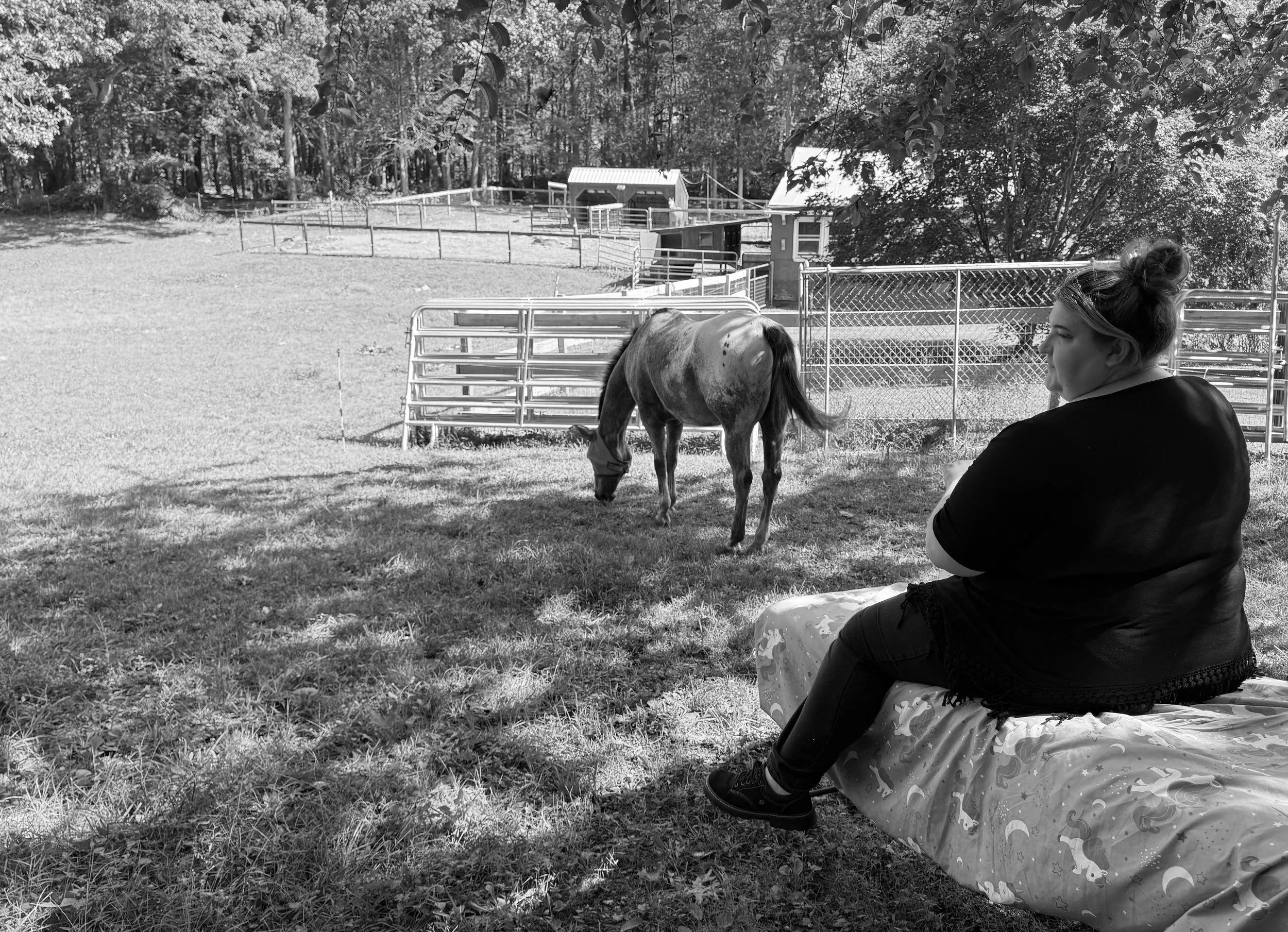 A woman sitting on a blanket with a horse grazing nearby in a fenced outdoor area. There are trees and a small building in the background.