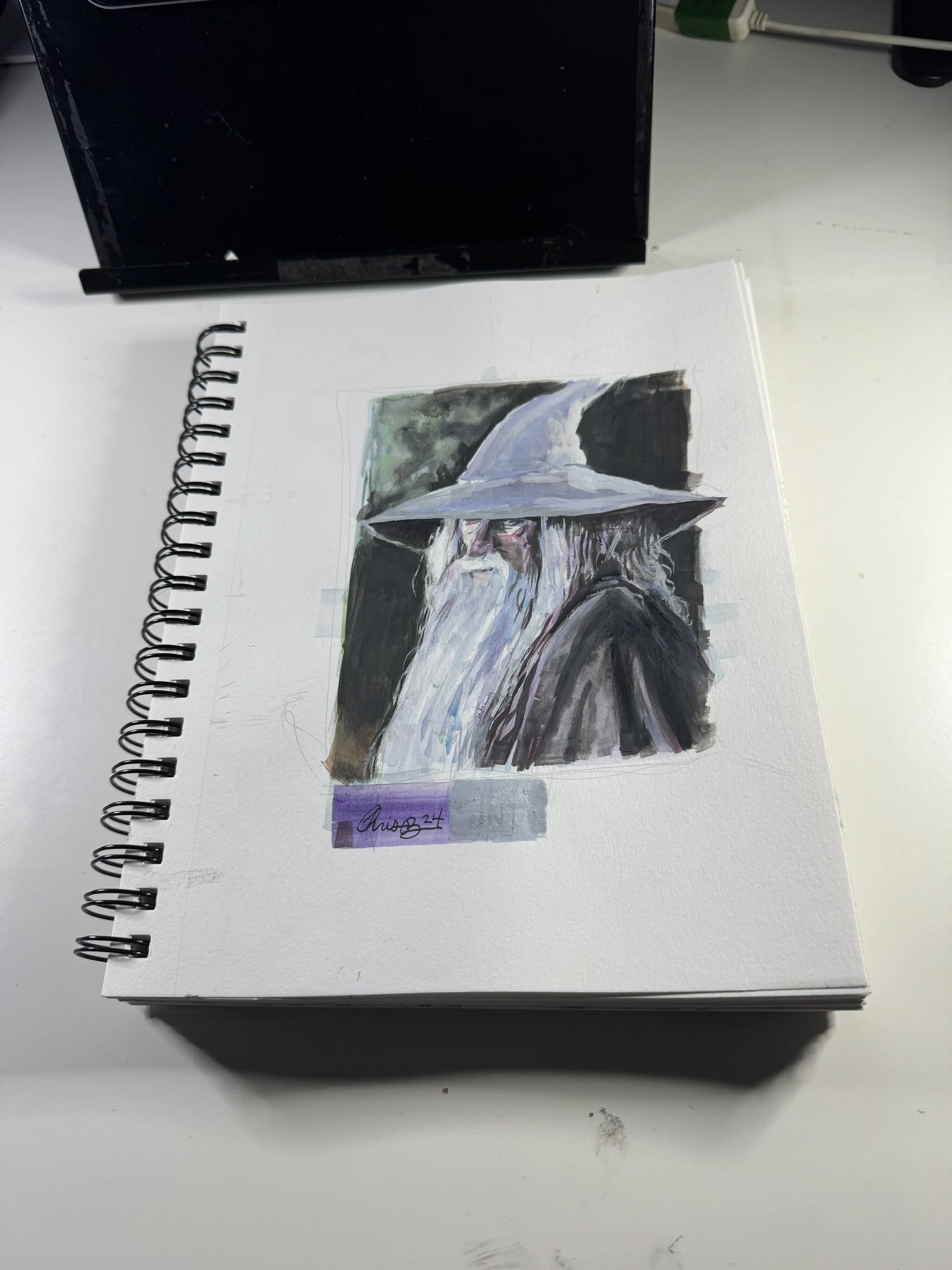 Watercolor painting of a wizard with a long white beard, wearing a tall pointed hat, set on a white sketchbook page on a white desk.