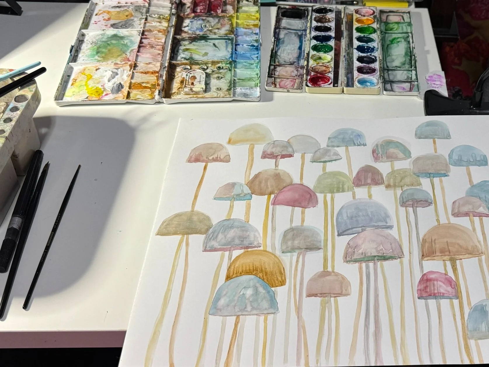 A watercolor painting of colorful mushrooms on a white sheet of paper, with watercolor paints and brushes visible on a table.