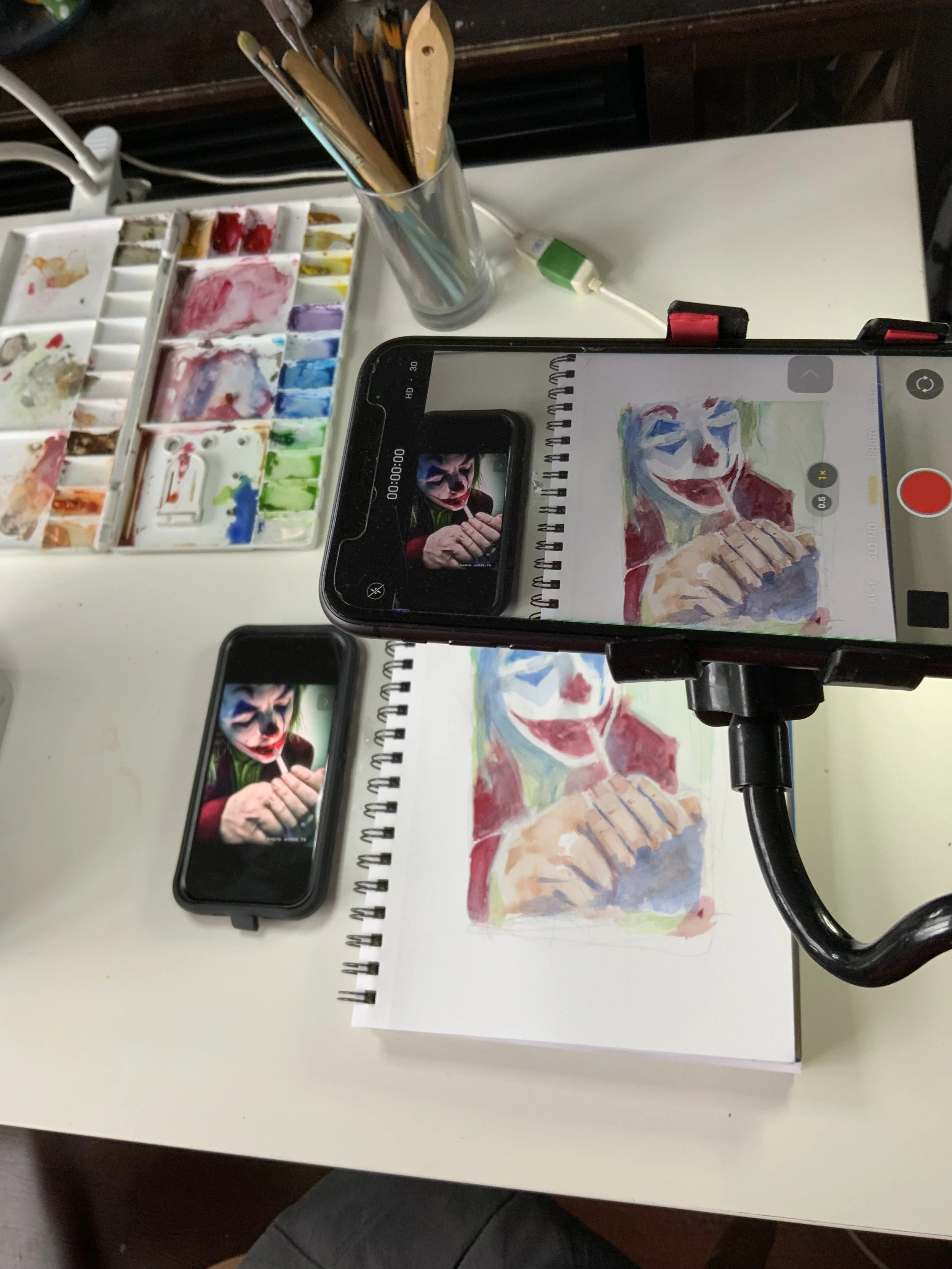 A watercolor painting of a clown with white face paint, red nose, and a red and green costume, depicting a person holding a cigar under a phone recording the artwork.