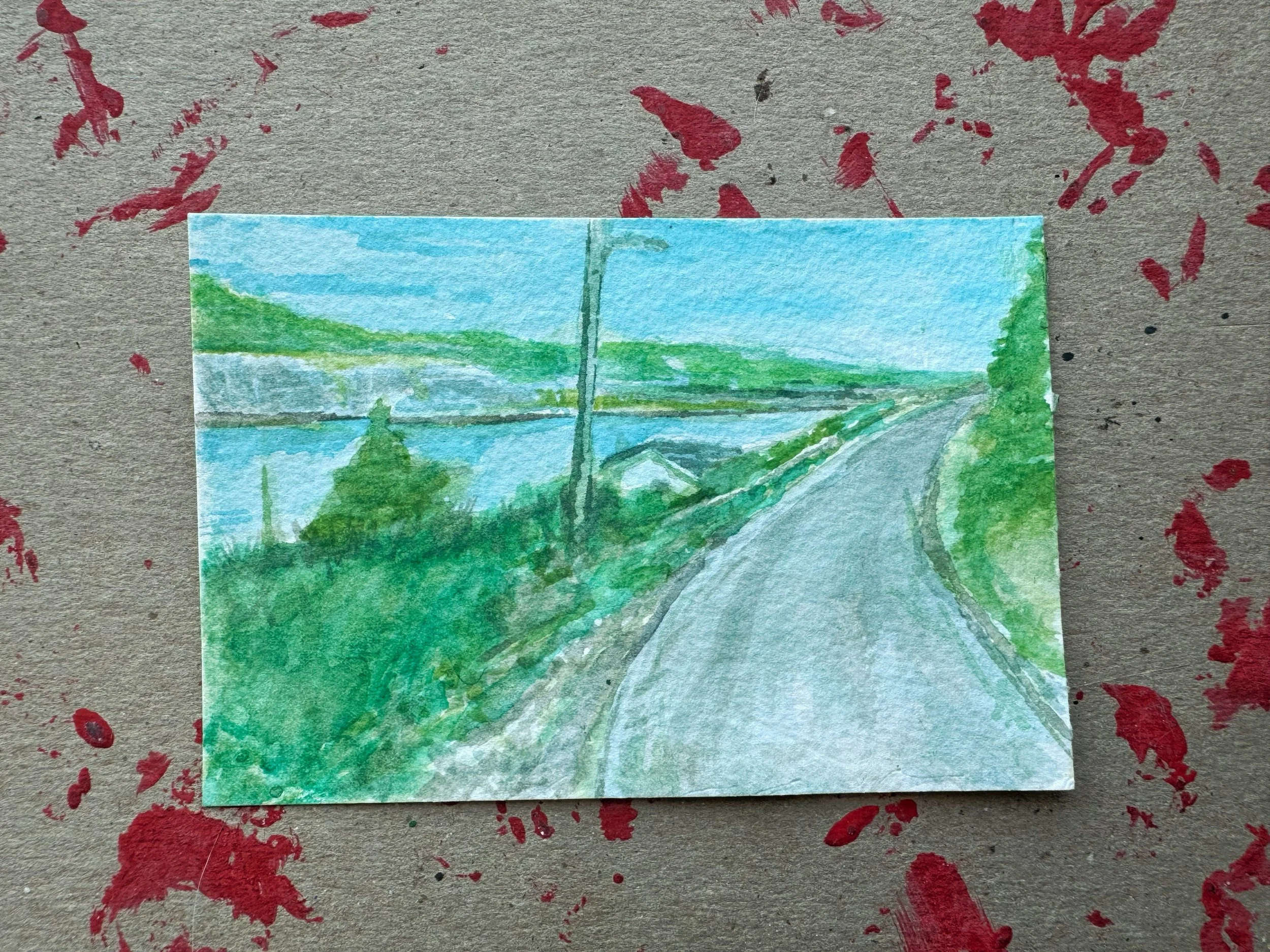 A small watercolor painting of a scenic landscape with a road, greenery, a body of water, and distant hills under a blue sky.