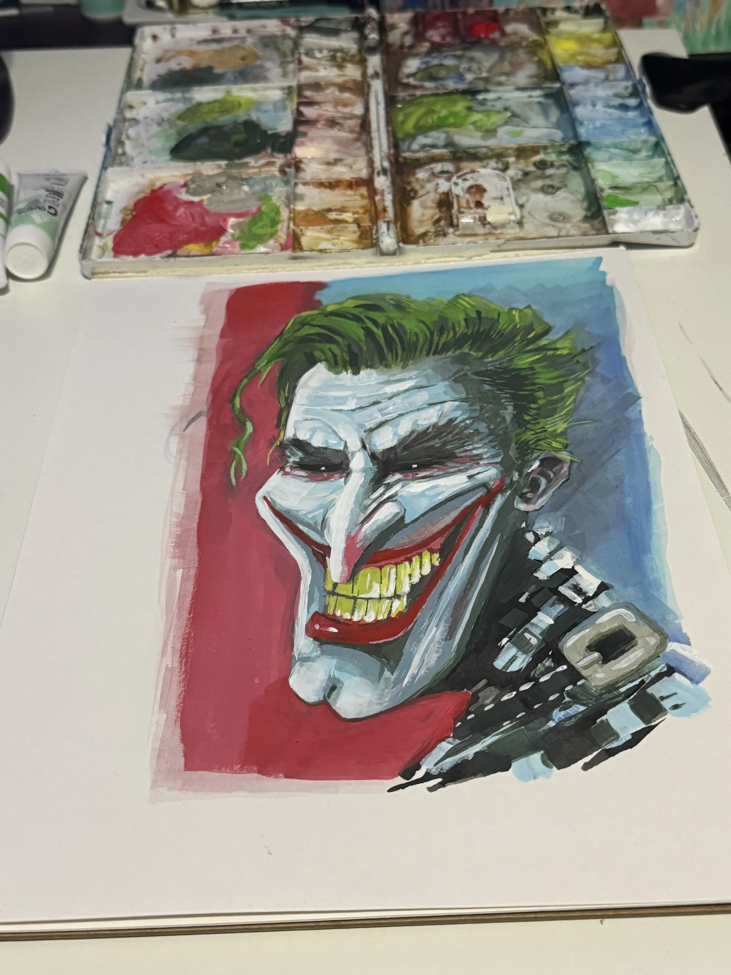 A colorful painting of the Joker from Batman, with green hair, white face paint, red lips, and a wide, sinister smile, on a piece of paper with an art palette and brushes in the background.