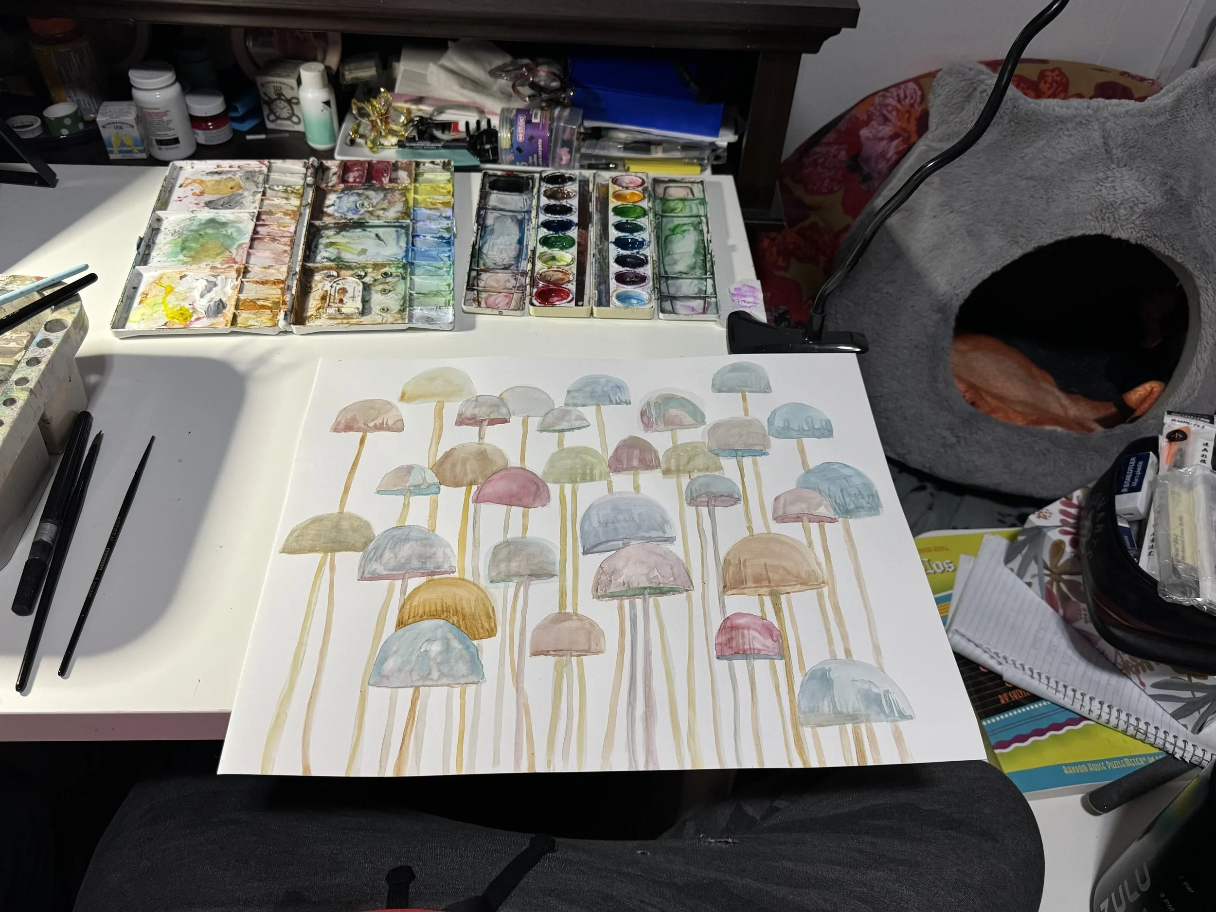 Art supplies on desk with watercolor painting of mushrooms in pastel colors.
