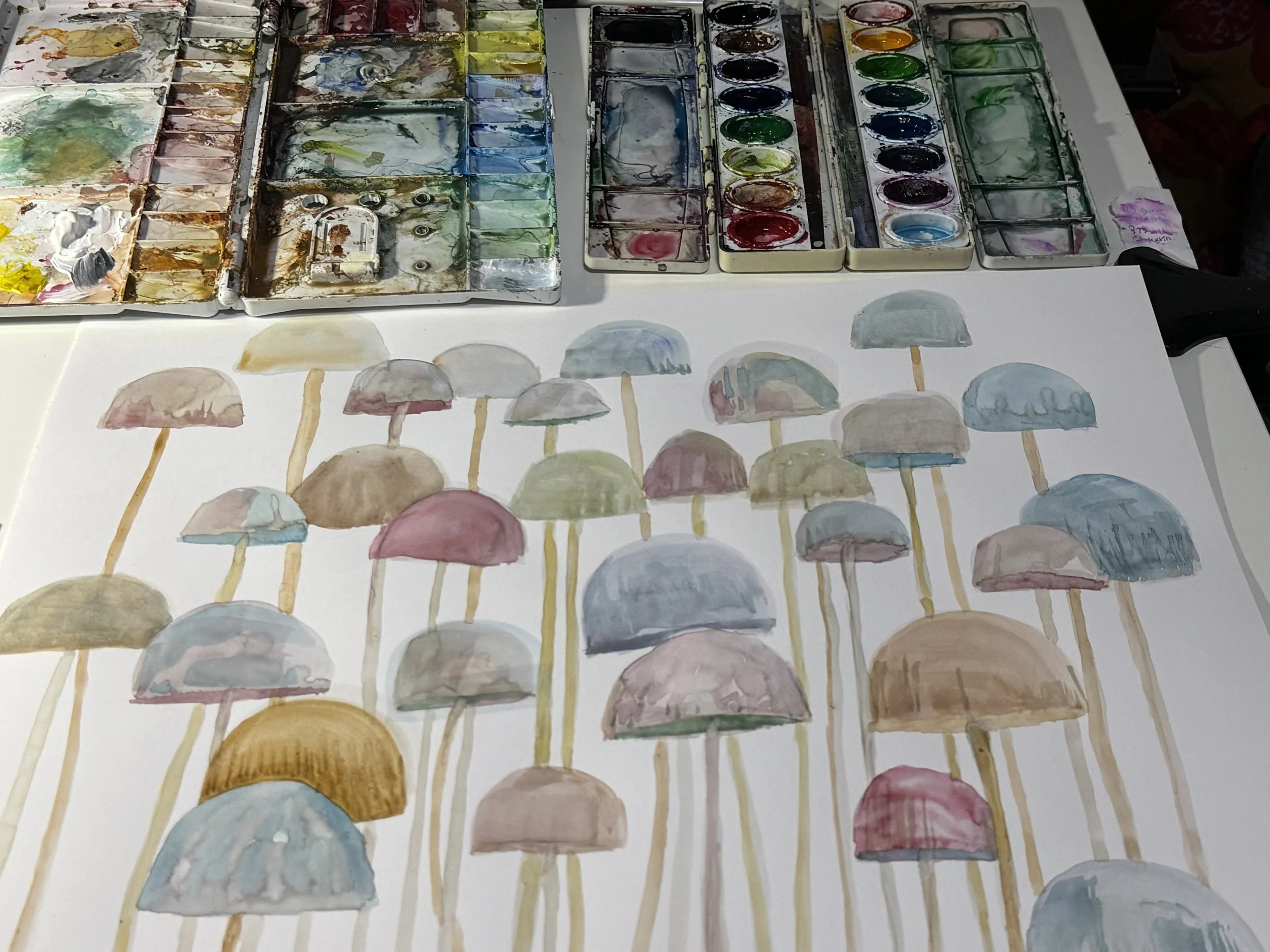 A watercolor painting of mushrooms in pastel colors, with a watercolor palette and brushes visible in the background.