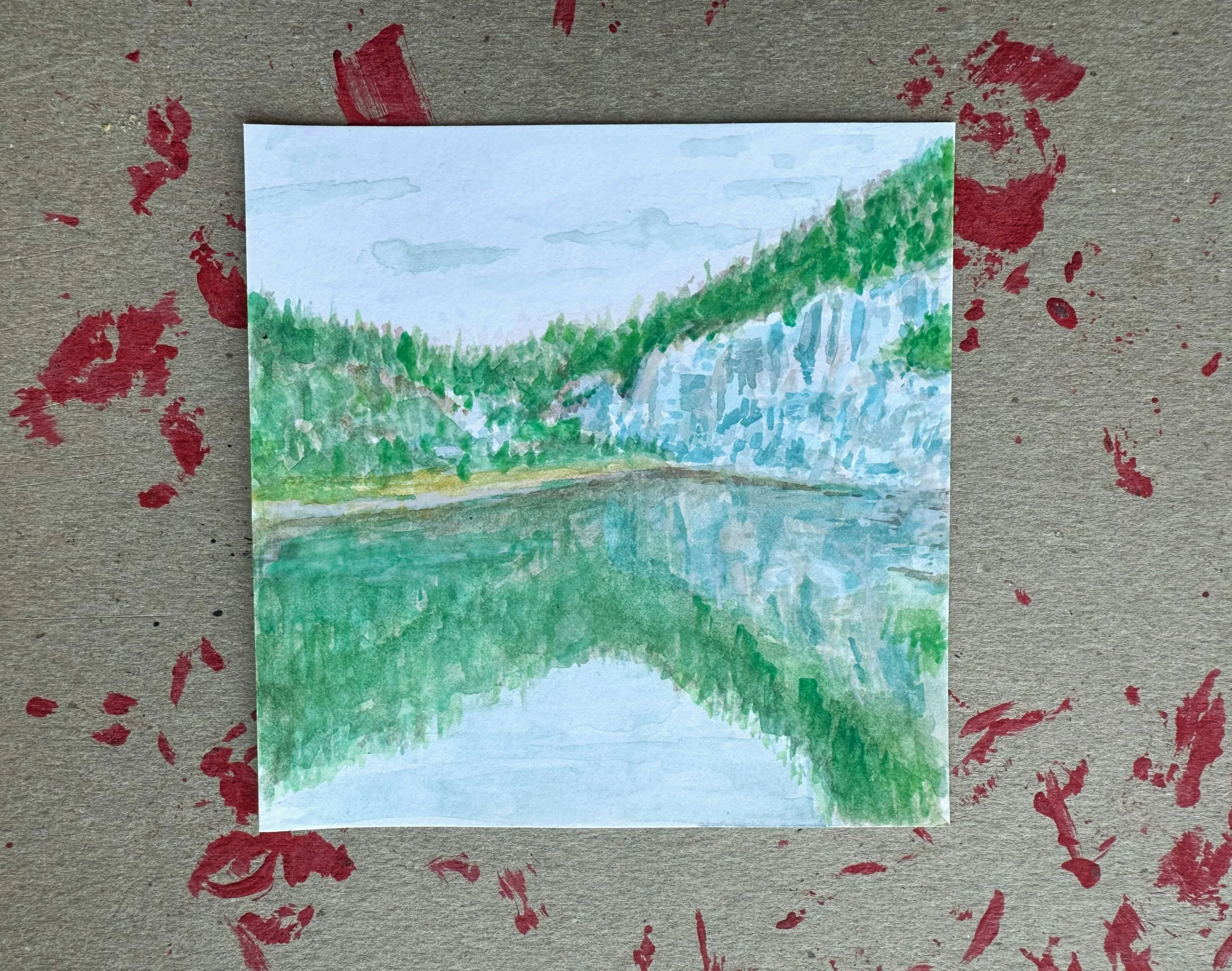 Watercolor painting of a landscape with a waterfall, green trees, and a pond reflecting the scenery, placed on a surface with red paint splatters.