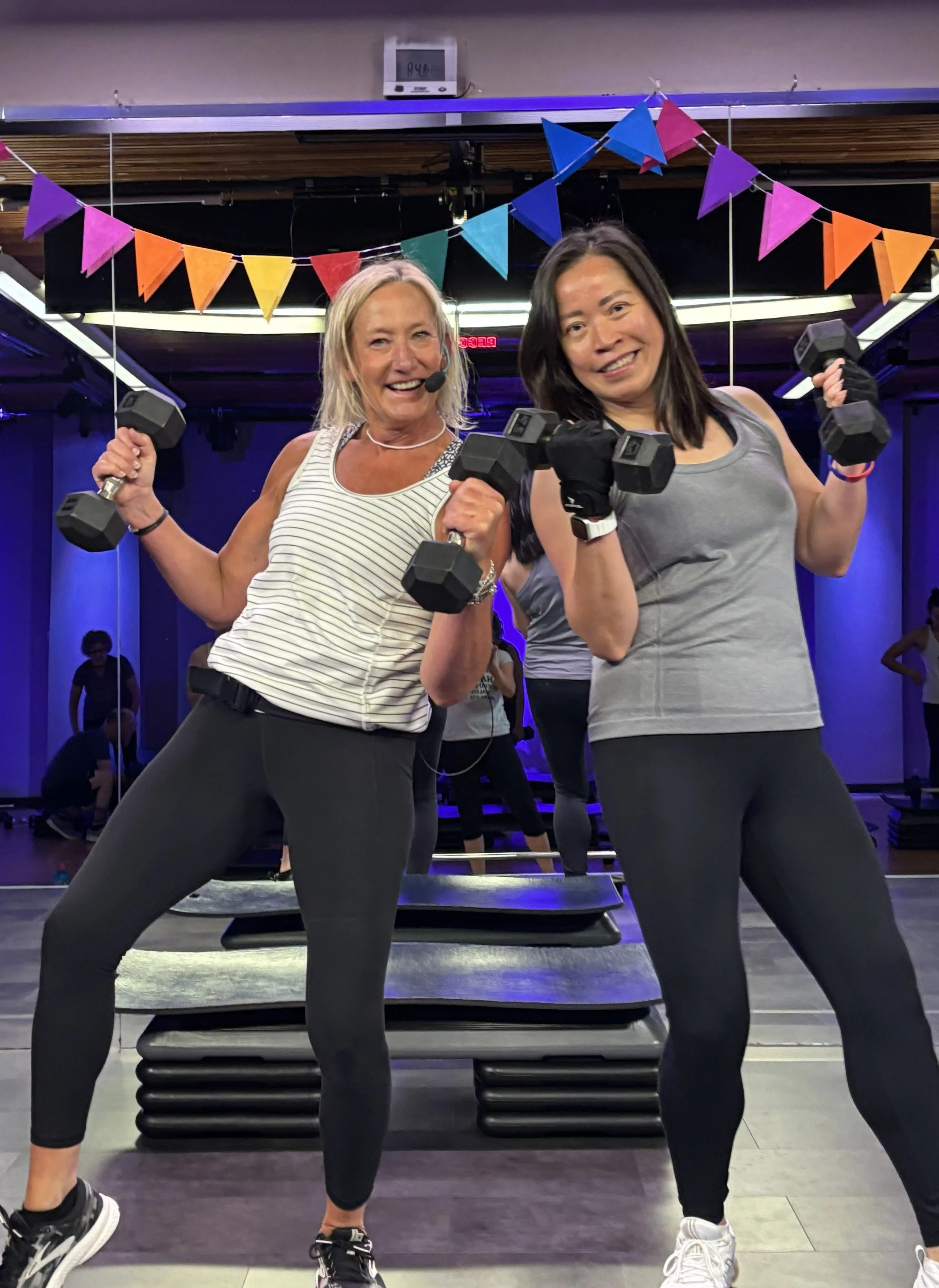 Two women smiling, holding dumbbells in a fitness class or gym with colorful banners overhead and exercise mats on the floor.