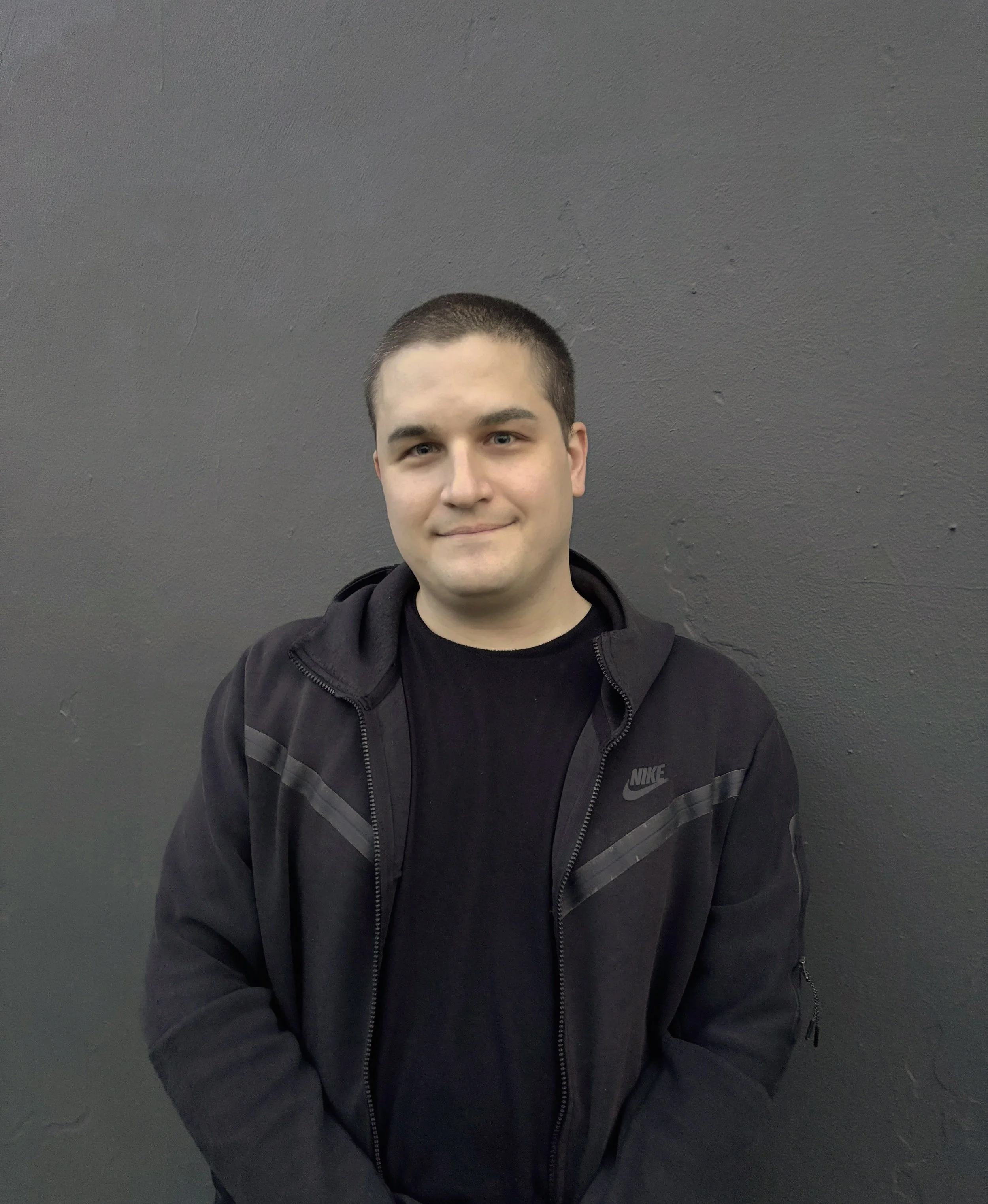 A young man with short dark hair wearing a black Nike jacket and black shirt standing against a dark gray wall.