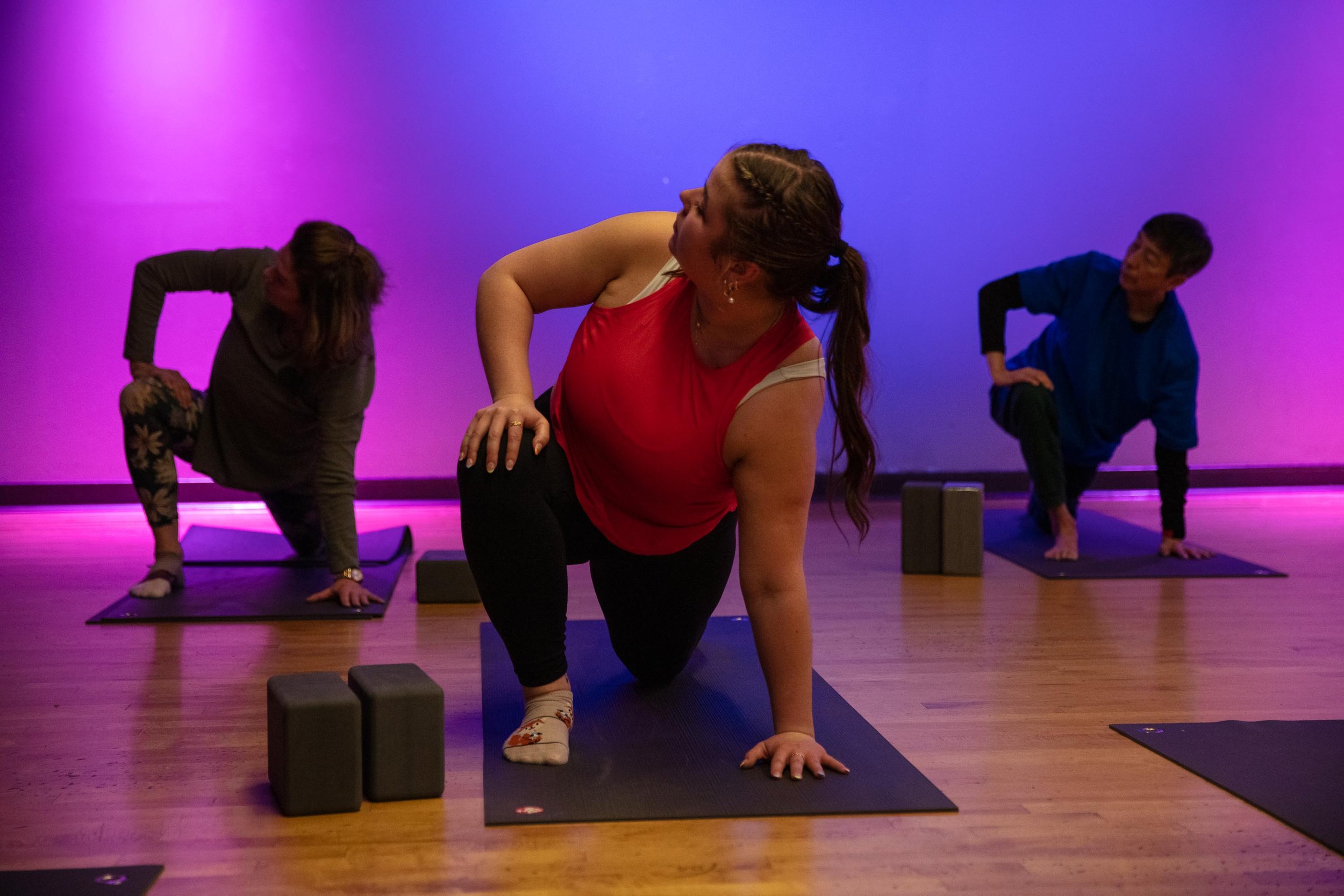 Group yoga class with participants in various poses on mats, dimly lit room with purple and blue lighting.