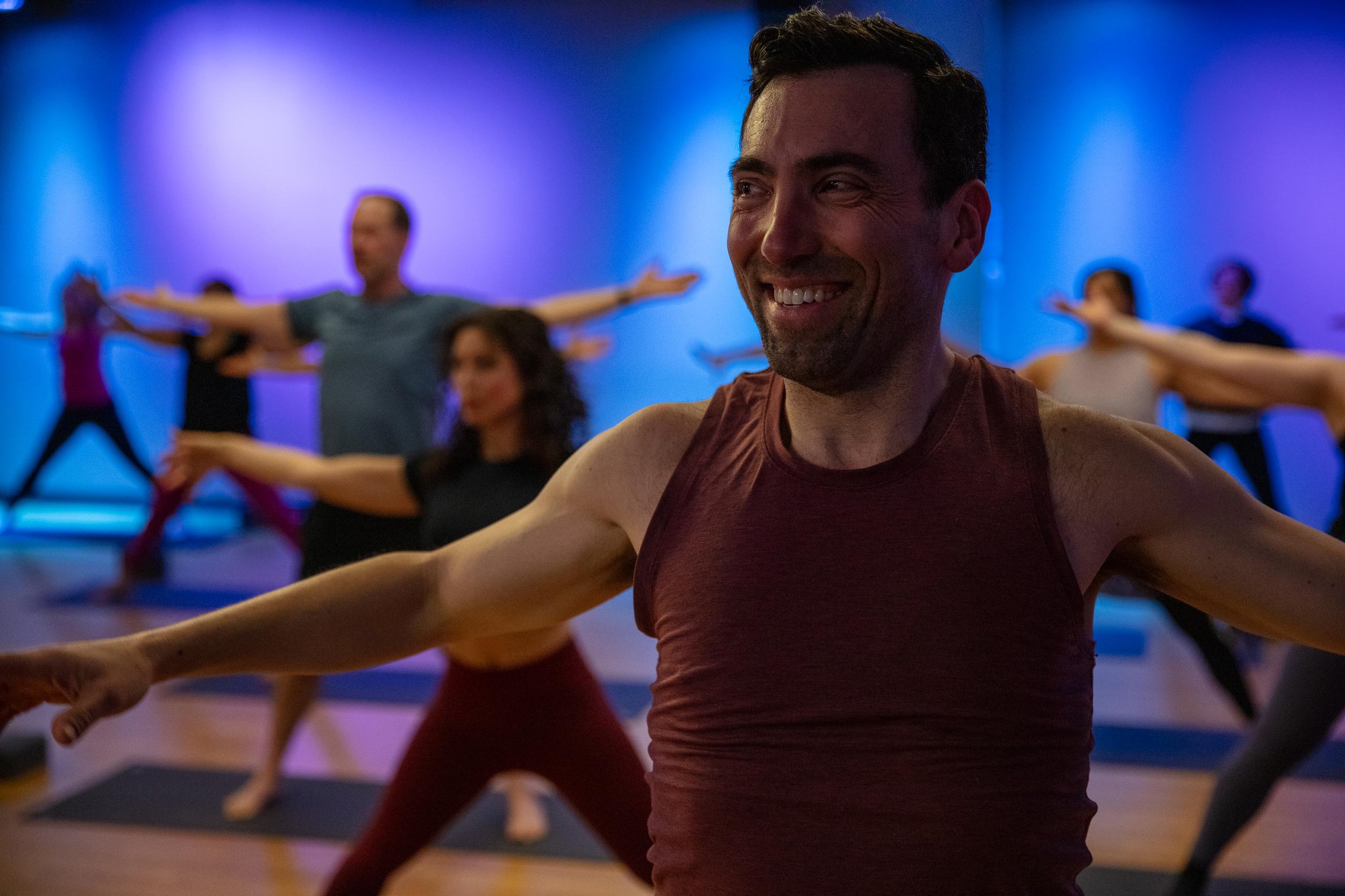 A group yoga class with people in a warrior pose, led by a man in a maroon tank top, in a dimly lit studio with purple and colorful lighting.