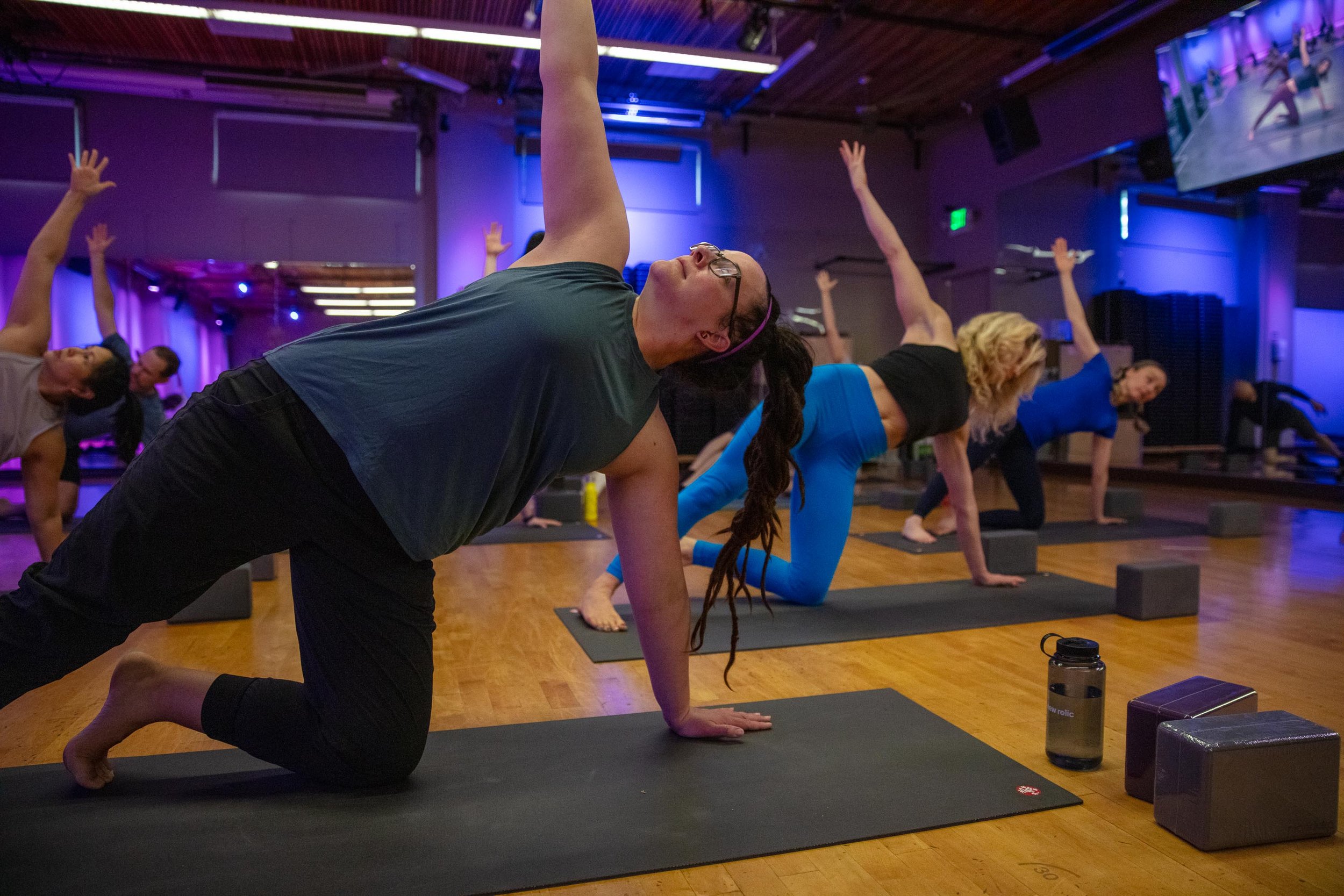 Group of people practicing yoga in a studio, performing a side stretch pose on mats with yoga blocks, under dim, colorful lighting.