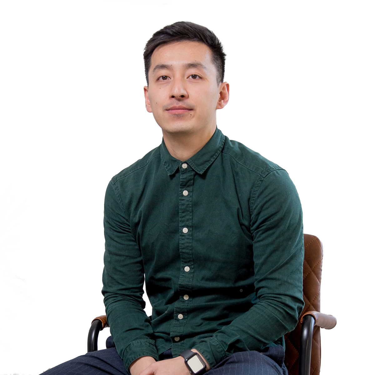 Portrait of a young man sitting on a chair, wearing a dark green button-up shirt and a smartwatch. He has short, dark hair and a neutral expression.