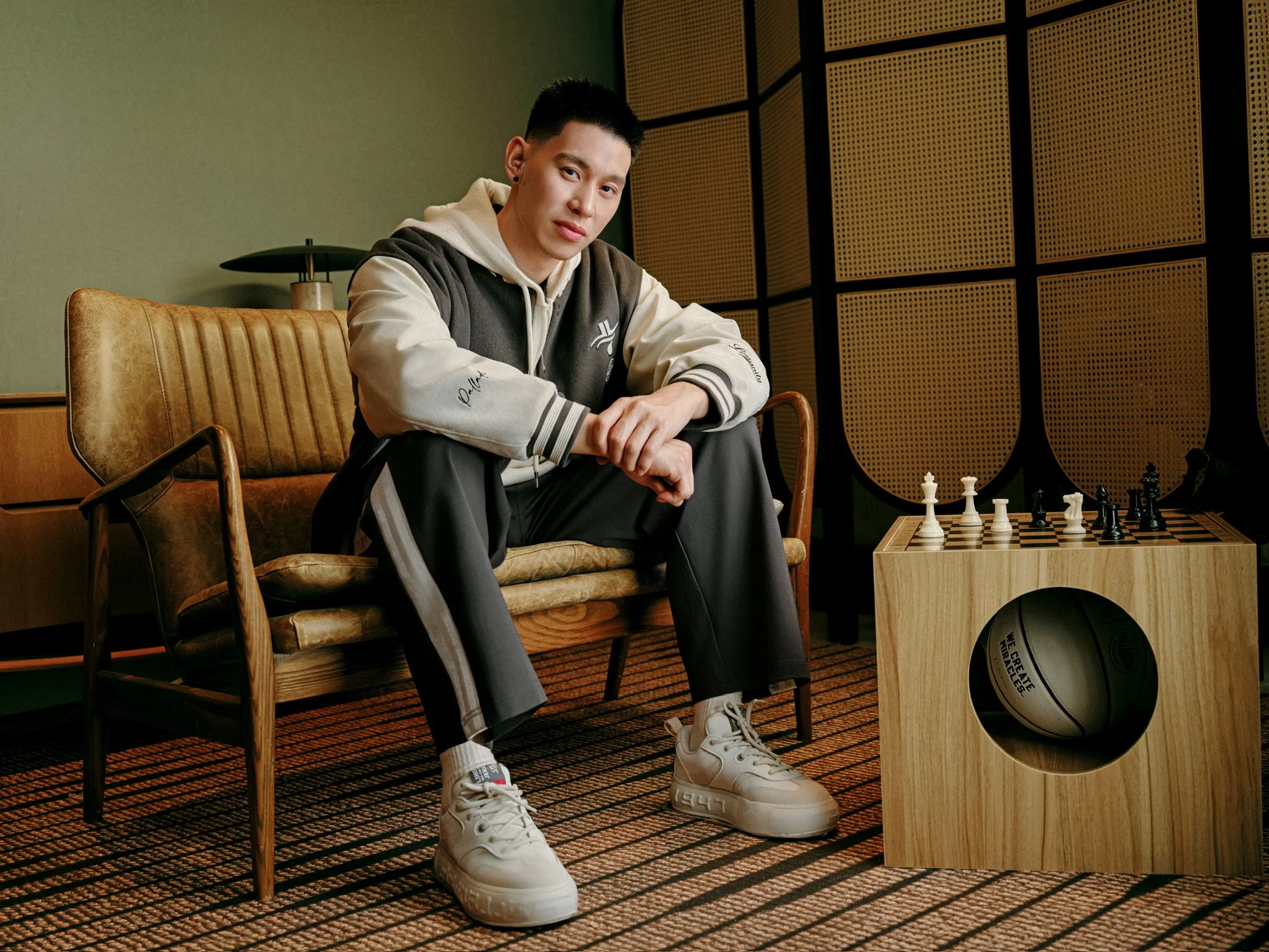 A young man sitting on a vintage-style leather chair in a room with a chessboard on a wooden table nearby. The room features acoustic wall panels, and the man is dressed in casual streetwear, wearing white sneakers, a hoodie, and a varsity jacket.
