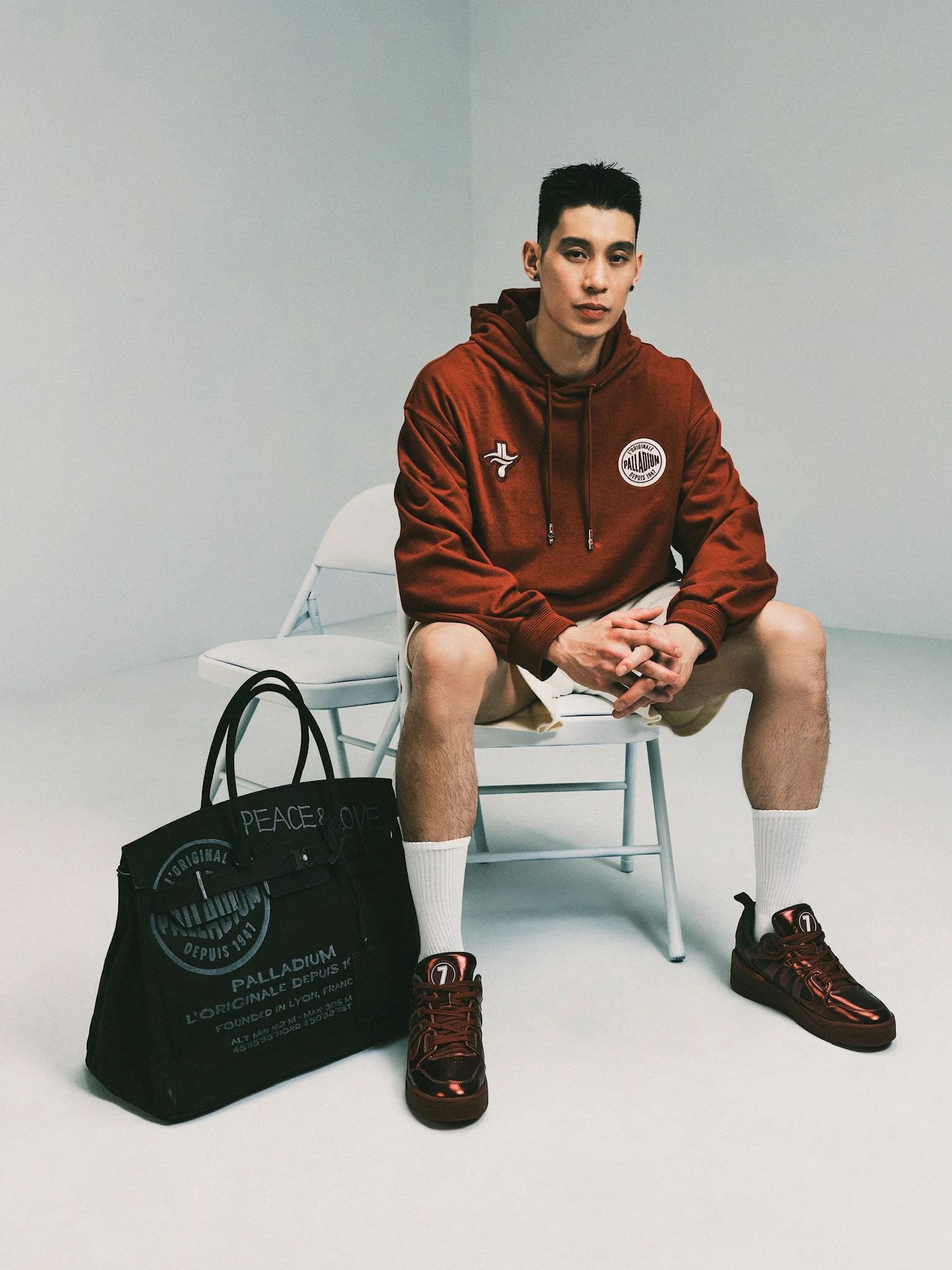 A young man sitting on a white chair in a minimalist room with light gray walls. He is wearing a red hoodie, beige shorts, white knee-high socks, and black sneakers with red accents. A black tote bag with white text is on the floor next to him.