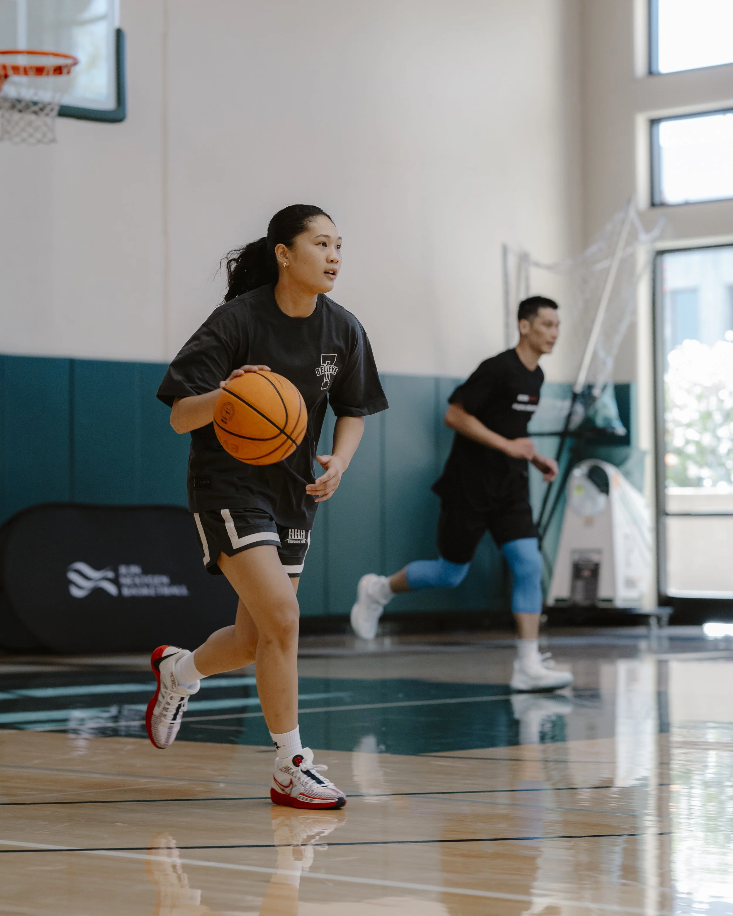A woman playing basketball on an indoor court with a man jogging in the background.