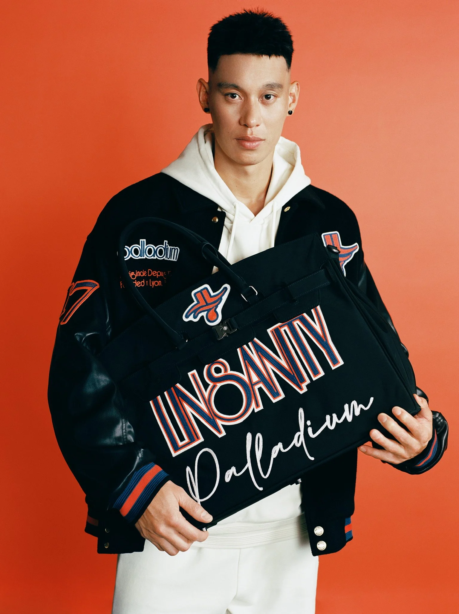 A young man with short black hair and earrings holds a black leather and fabric bag with colorful text that reads 'UNSONATY Palladium'. He wears a black and orange varsity jacket over a white hoodie and stands against a reddish-orange background.