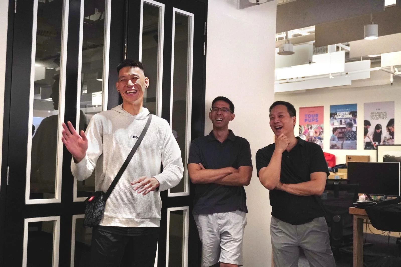 Three men standing indoors, laughing and smiling, with posters on the wall behind them.