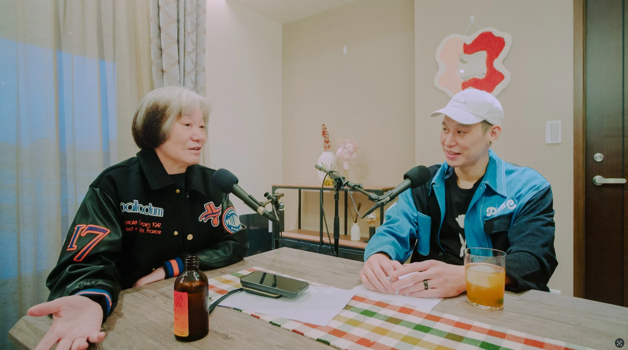 Two people sit at a table with microphones, engaged in conversation. The person on the left is older with short gray hair, wearing a black jacket with patches, and gesturing with one hand. The person on the right is younger with short hair, wearing a white cap, a blue jacket, and a black shirt, smiling. On the table are a bottle, a phone, a glass of orange drink, and some papers. The background includes a wall with a floral arrangement, a small table, and a wall decor piece.