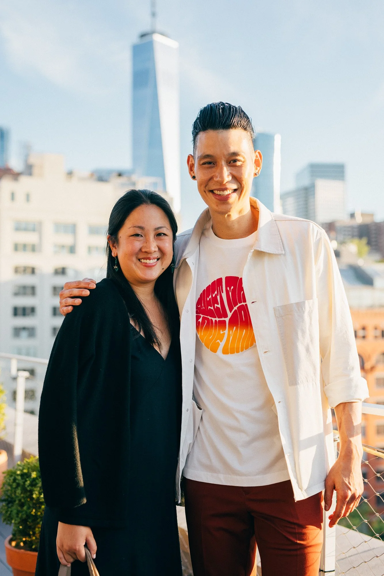 Smiling woman and man standing on a balcony with city skyline in the background during the day.