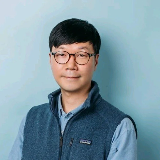 A man with black hair and glasses wearing a blue shirt and a blue zip-up vest, standing against a light blue background.