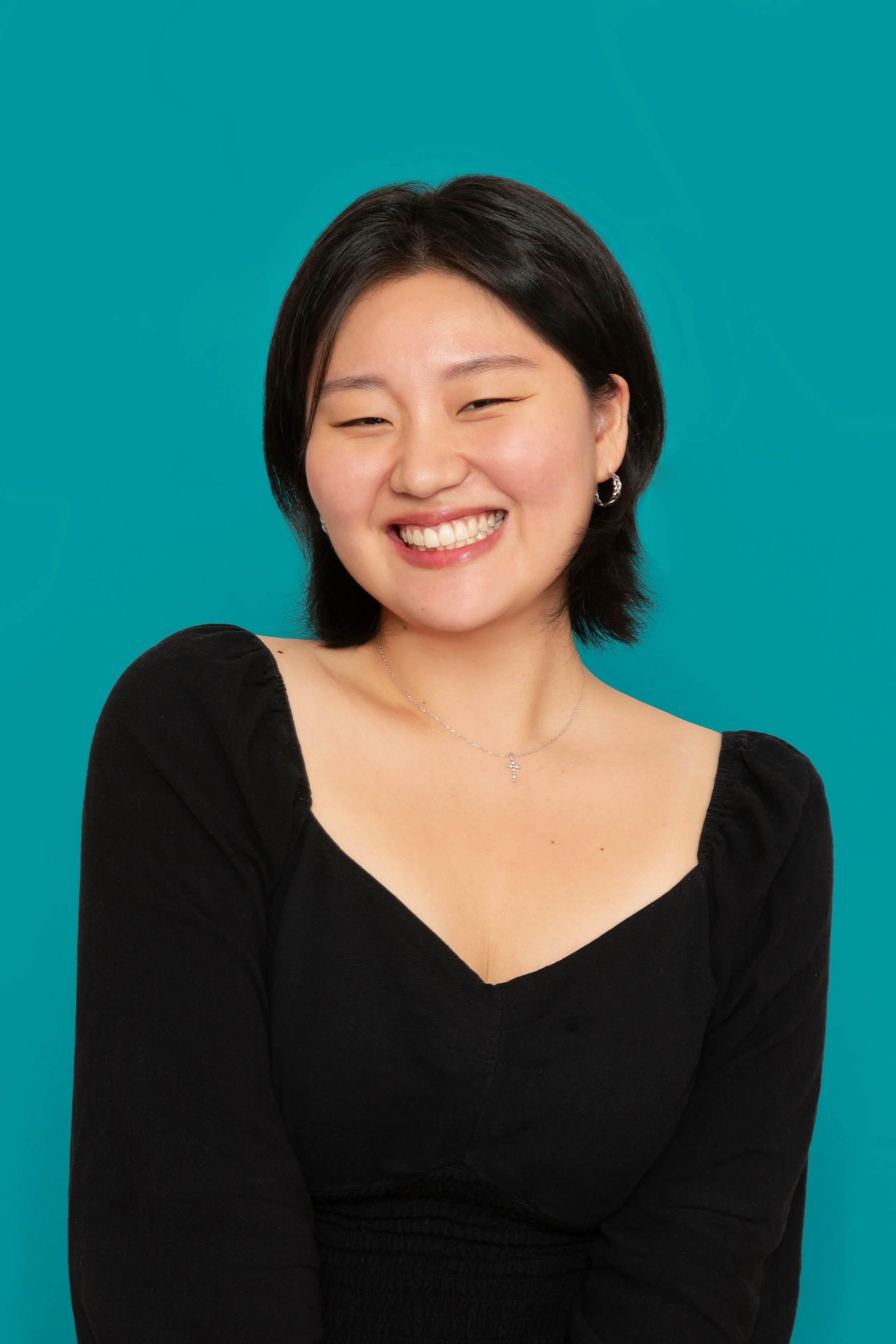 A young Asian woman with short black hair smiling in front of a teal background, wearing a black top, a silver cross necklace, and hoop earrings.